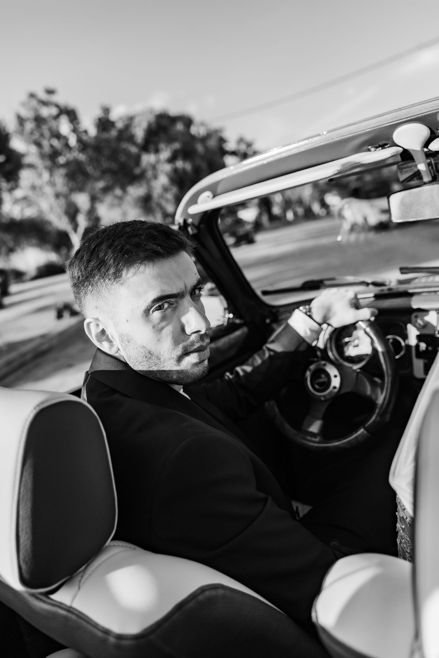 Black and white photo of a stylish man in a suit sitting in a vintage convertible car, looking over his shoulder.