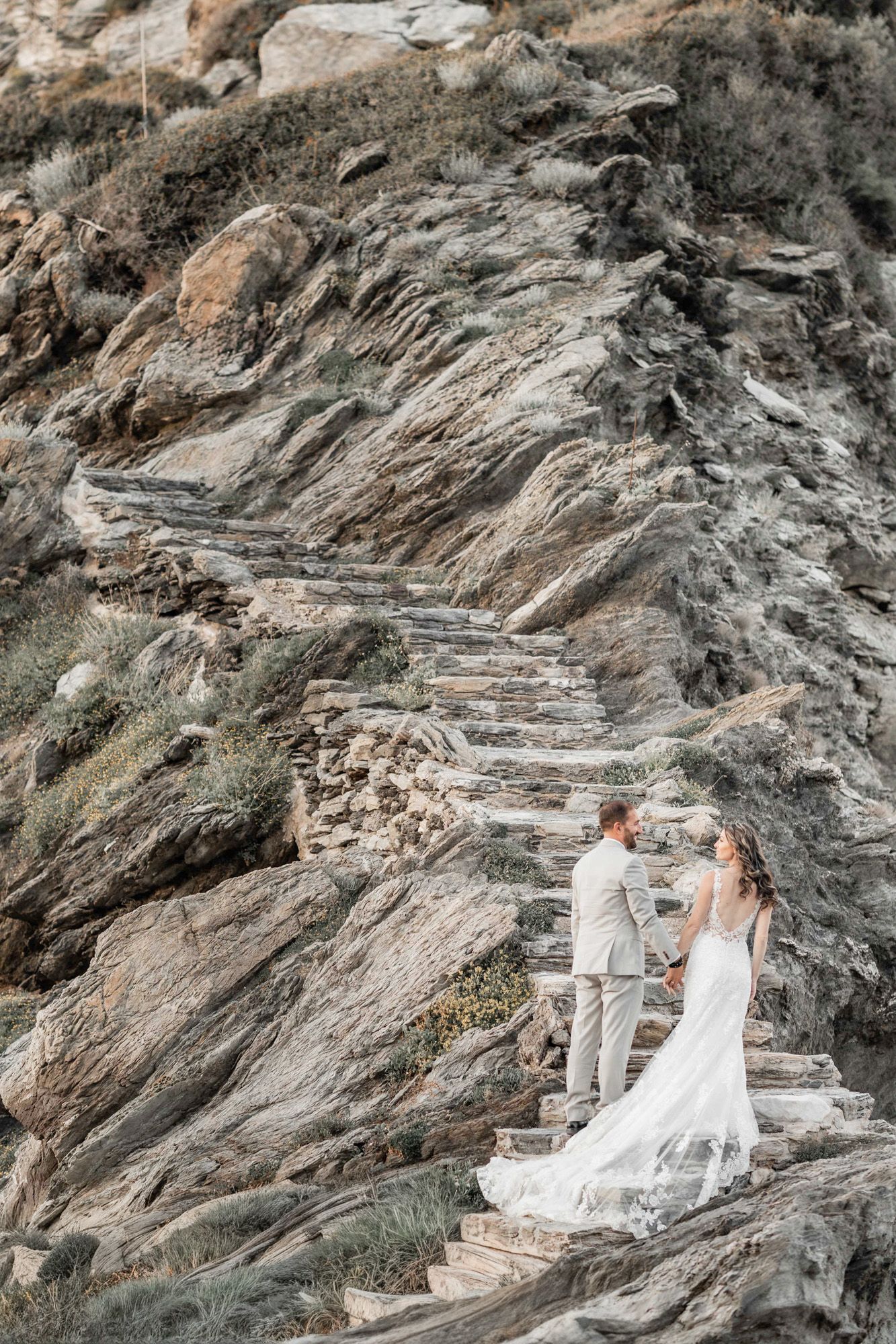 A couple in wedding attire stands on stone steps carved into a dramatic rocky cliff face.