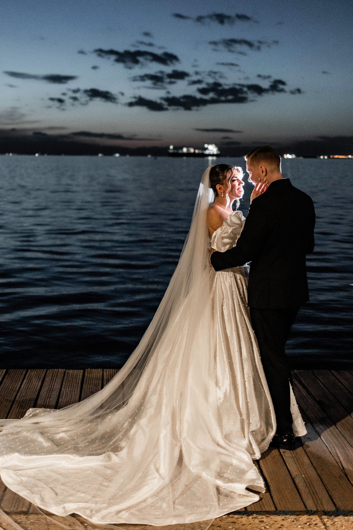 A romantic wedding couple embraces on a dock at twilight with a stunning waterfront view and dramatic sky.