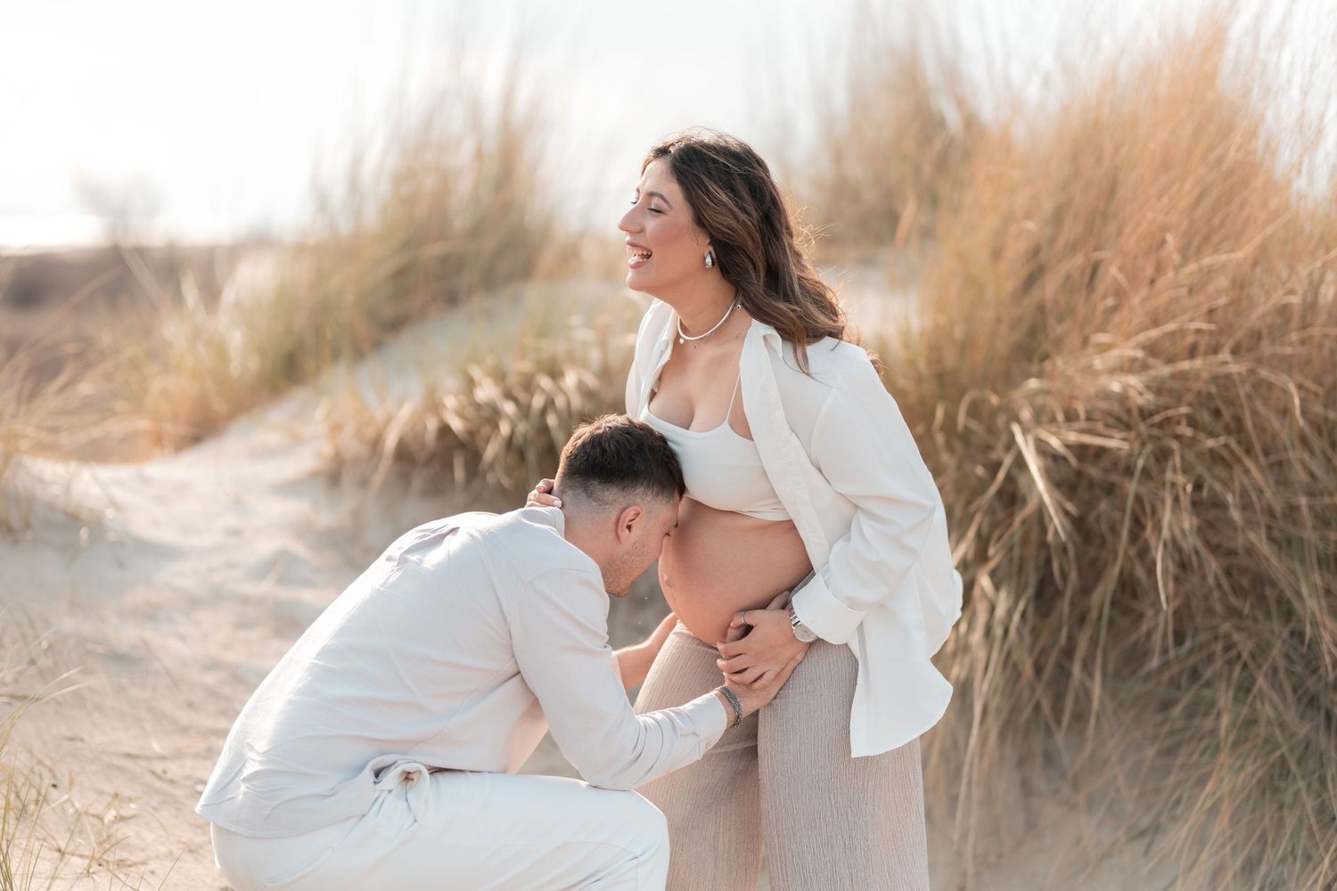 Romantic couple embraces on sandy beach at sunset with golden light and beach grass in background.