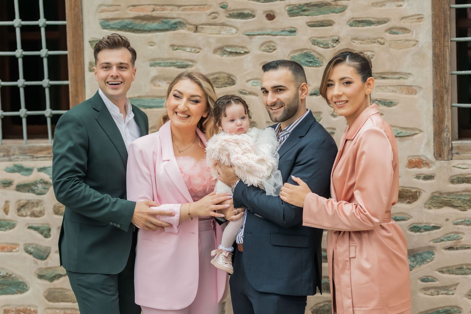 A group in coordinating formal attire poses together against a stone wall, with pink blazers and dark suits.