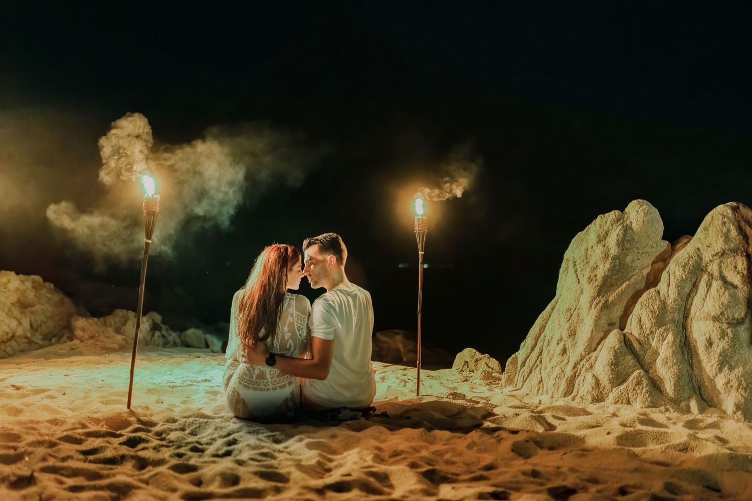 Romantic couple sits on sandy beach at night surrounded by glowing torches and dramatic rock formations.