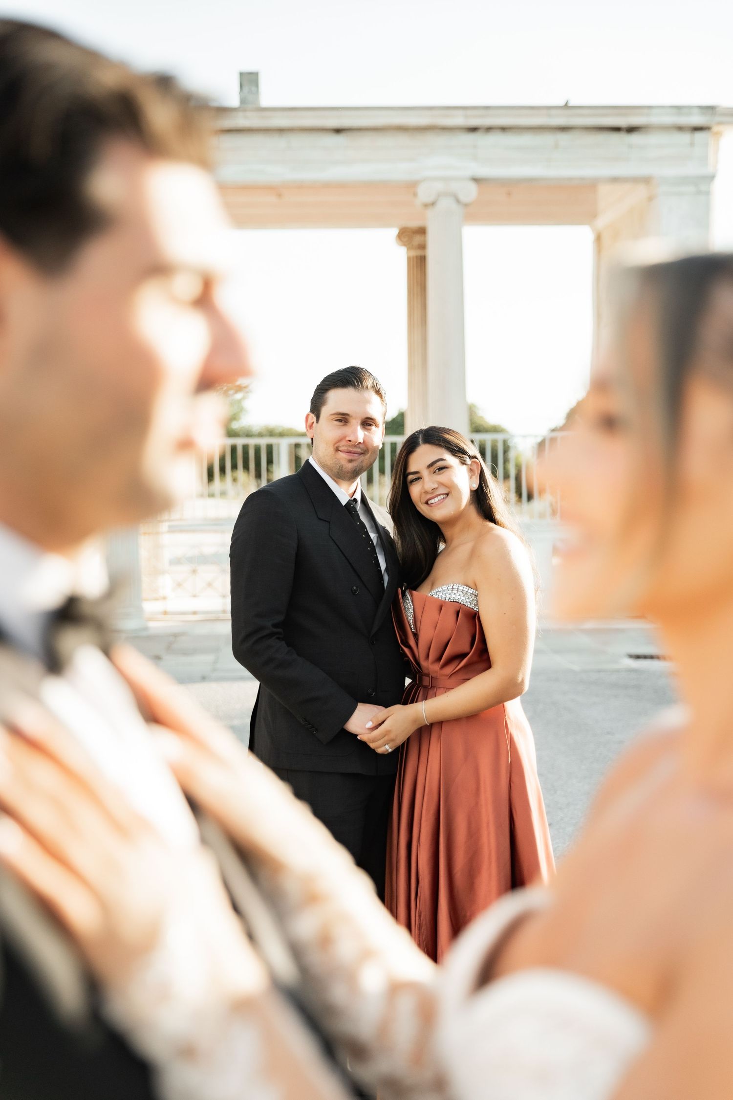 Couple in formal attire posing at an outdoor venue with white columns, man in black suit, woman in rust strapless gown.