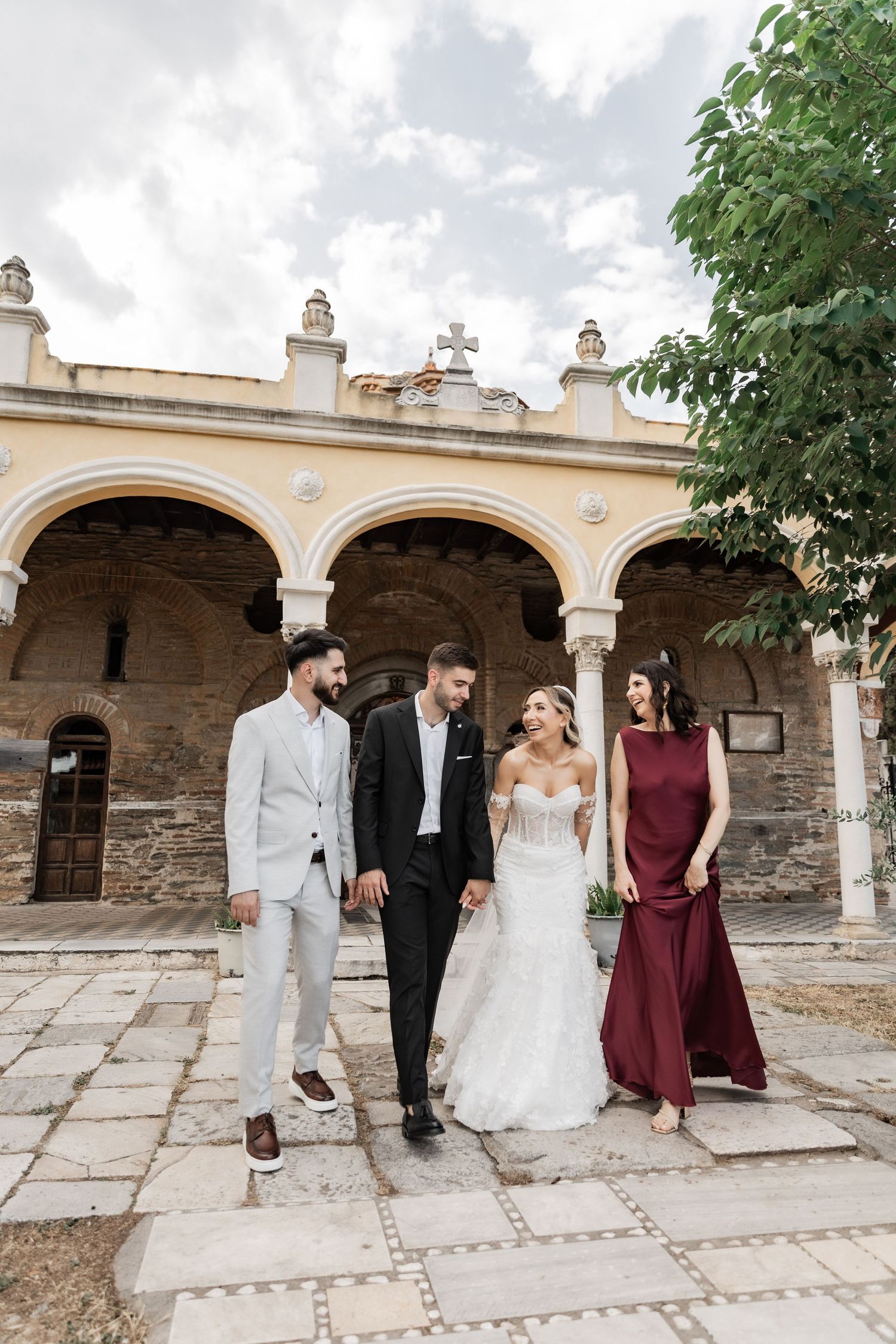 Bride and groom walk with wedding party outside historic stone church with arched colonnade on cloudy day.