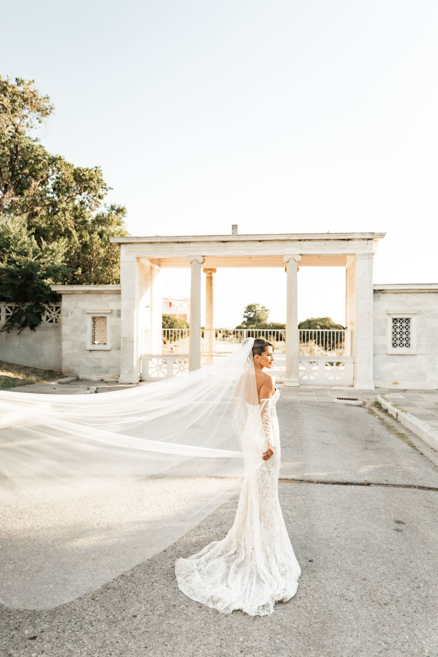 Bride in lace gown with flowing veil stands before elegant white colonnade at golden hour wedding venue.
