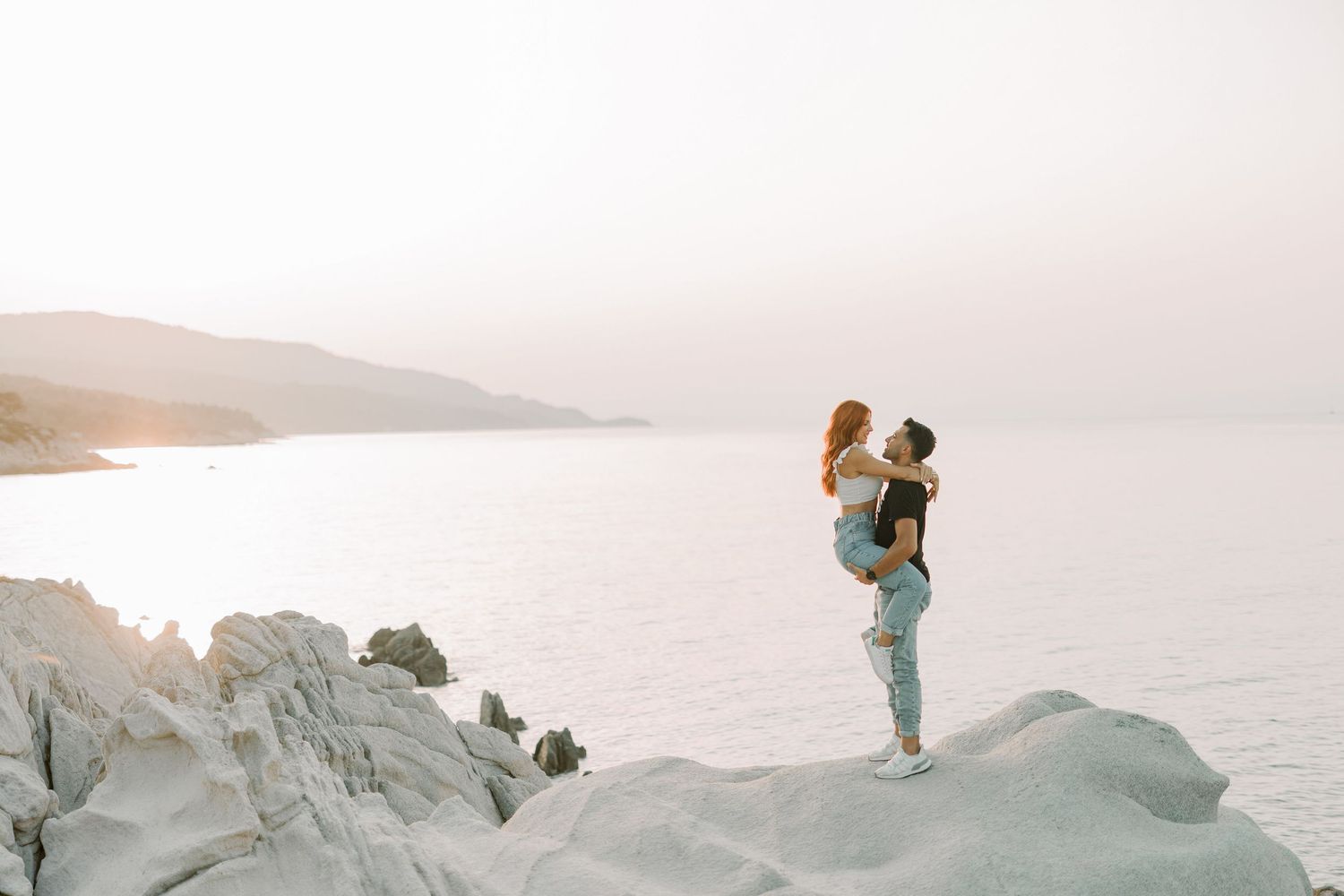 A couple embraces on a snowy mountain peak at sunset with dramatic winter landscape views in the background.