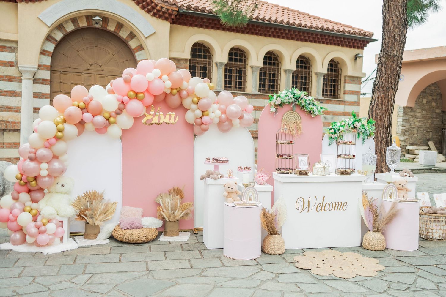 Pink and peach balloon garlands decorate an elegant outdoor party venue with Mediterranean-style architecture.