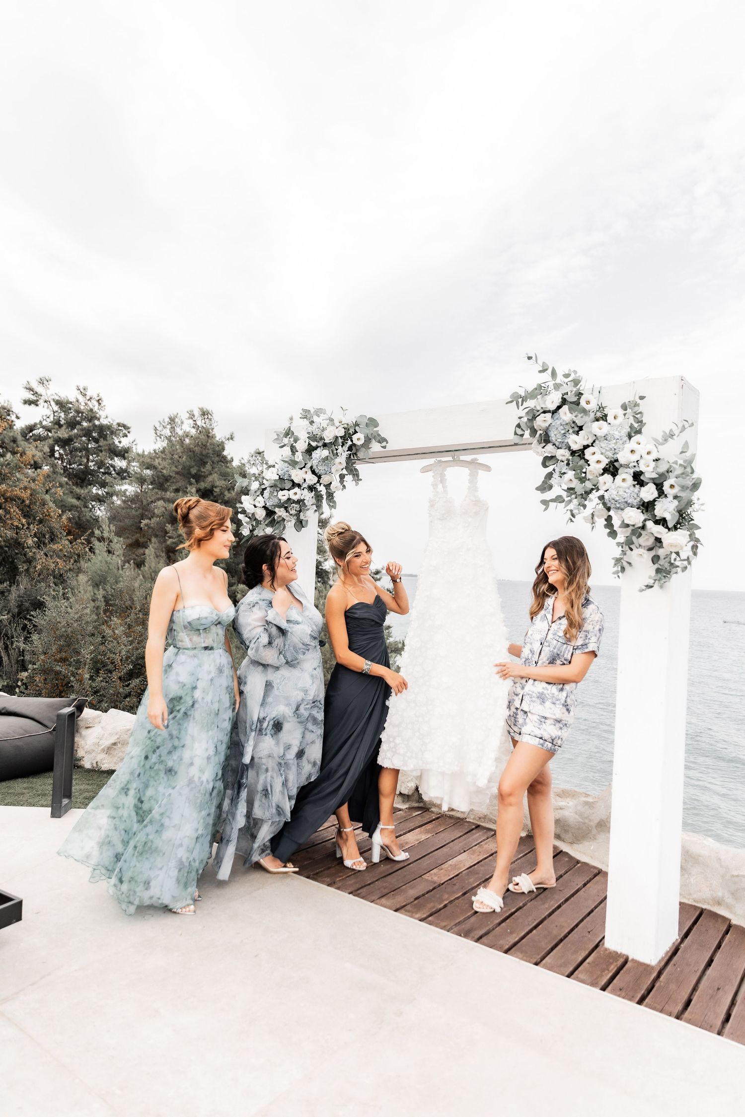 Wedding party in flowing blue dresses posing on a seaside deck with white pillars.