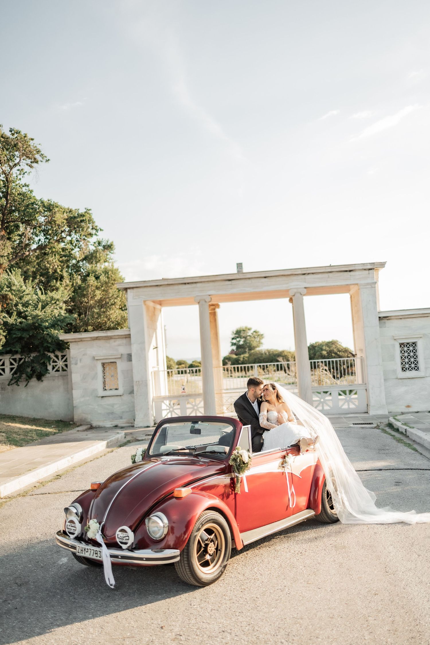 Bride and groom posing with a vintage red VW Beetle convertible decorated with flowers outside a classical building.