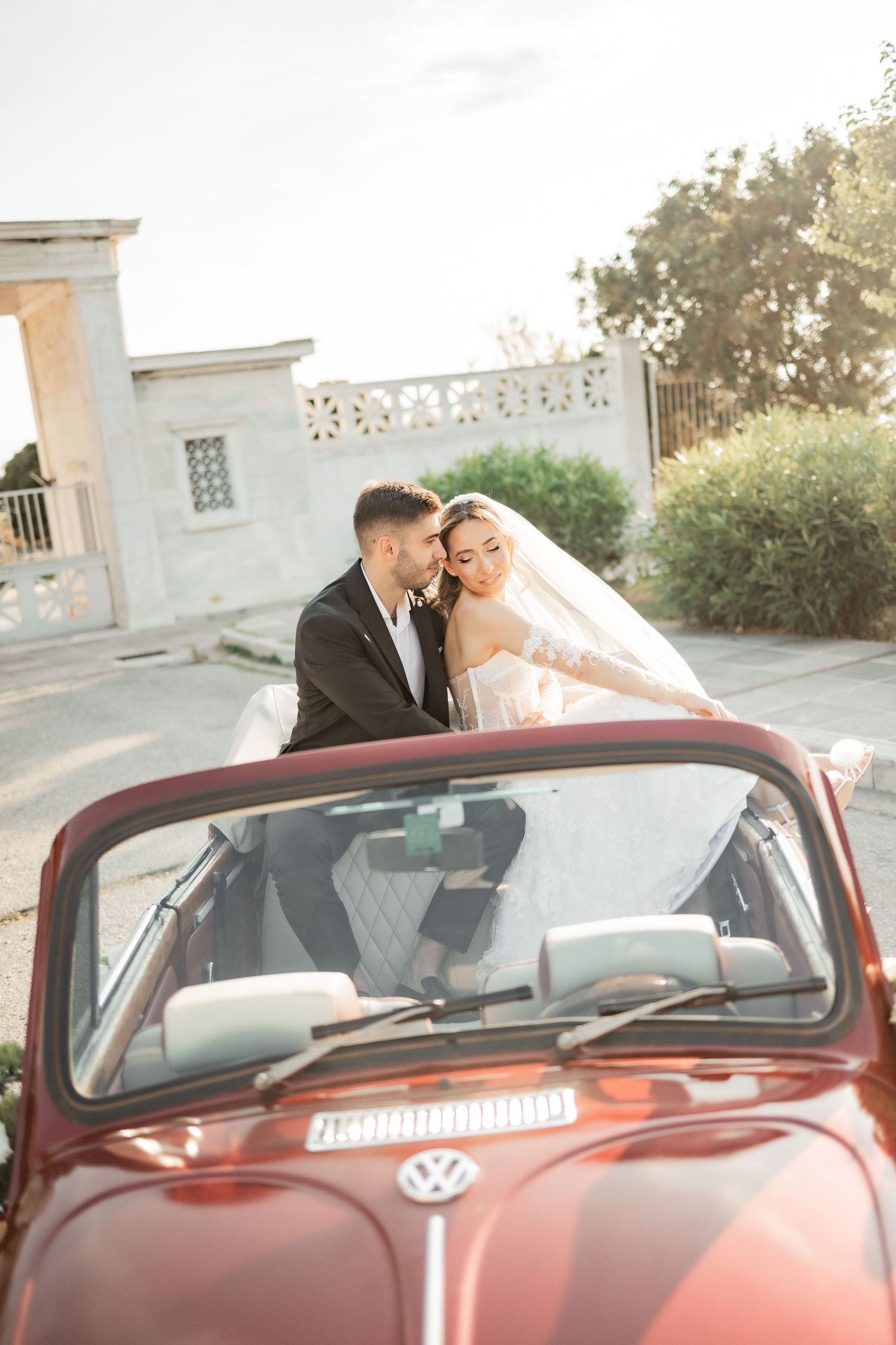 Newlywed couple sharing a romantic moment beside a vintage red VW Beetle convertible on their wedding day.
