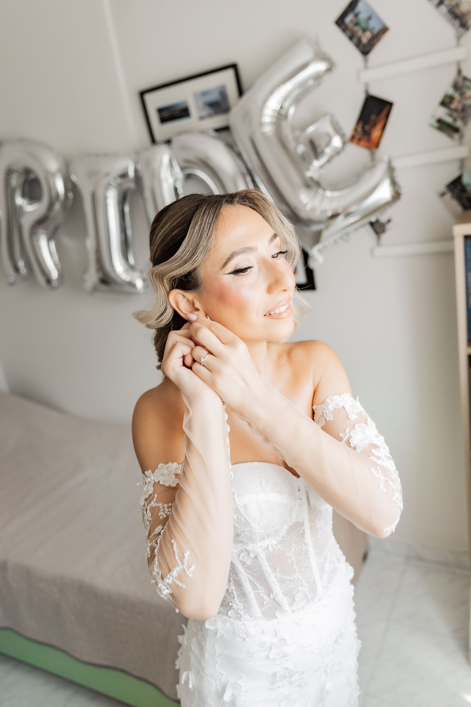 Bride in white lace corset gown putting on earrings with silver BRIDE balloons in background.