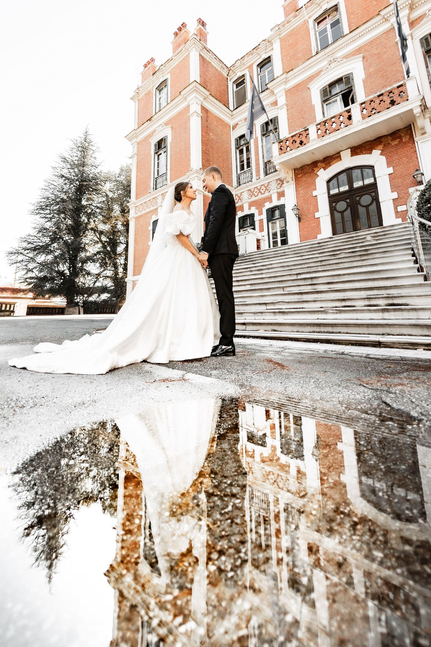 Wedding couple stands on mansion steps with their reflection in a puddle creating a mirror image.
