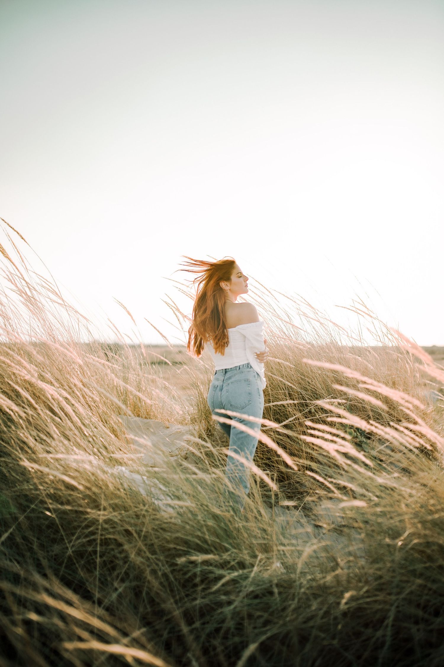 A person in a light sweater stands among tall windswept grass at sunset, creating a dreamy silhouette against the sky.
