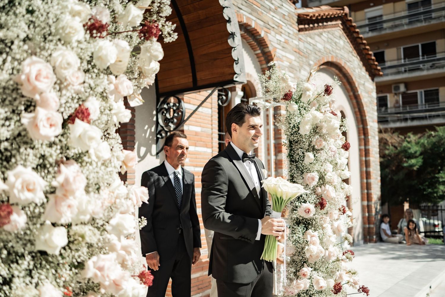 Groom in black tuxedo holds white bouquet beside floral arch of roses at elegant outdoor wedding ceremony.