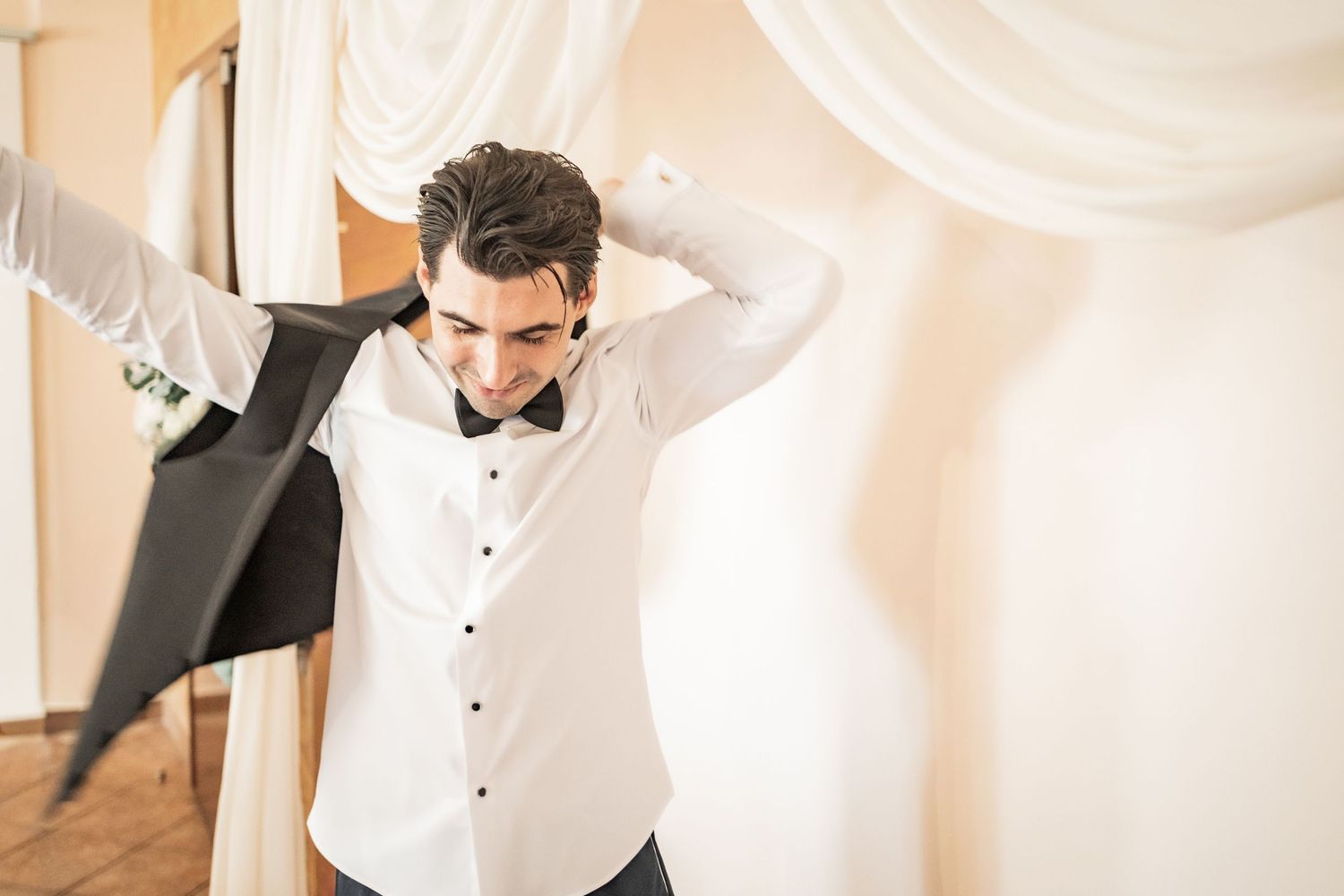 Young groom in white shirt and bow tie putting on suit jacket before wedding ceremony.