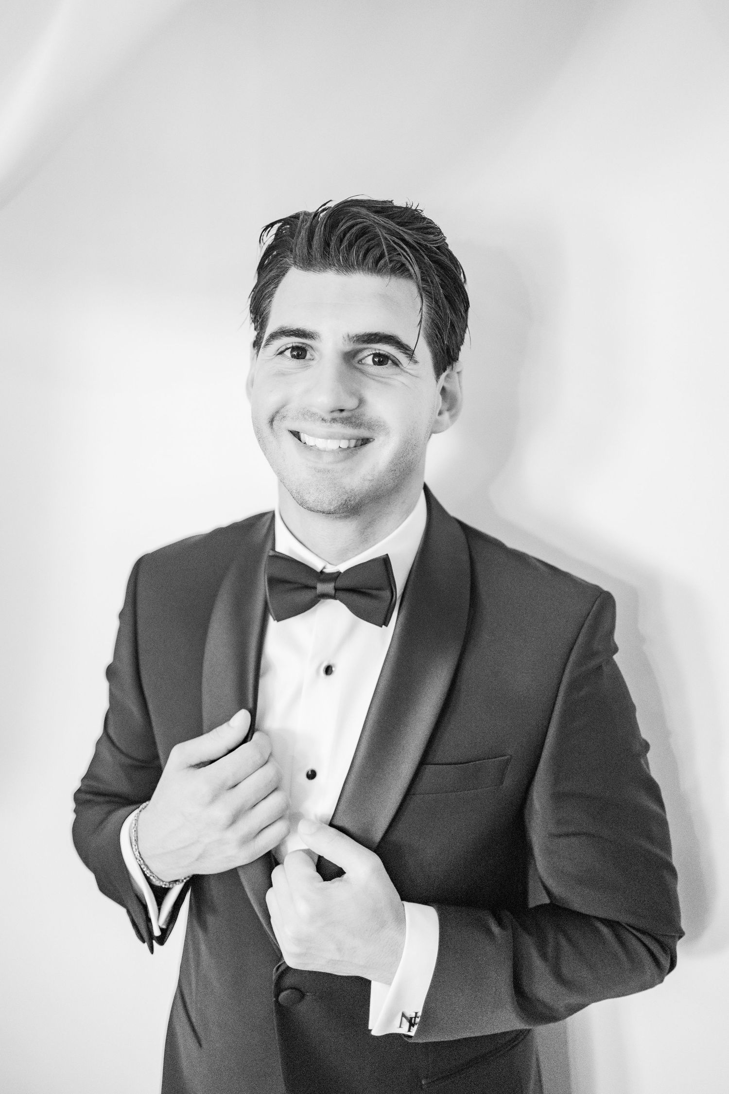 Smiling young man in a tuxedo with bow tie adjusting his jacket, black and white formal portrait photo.