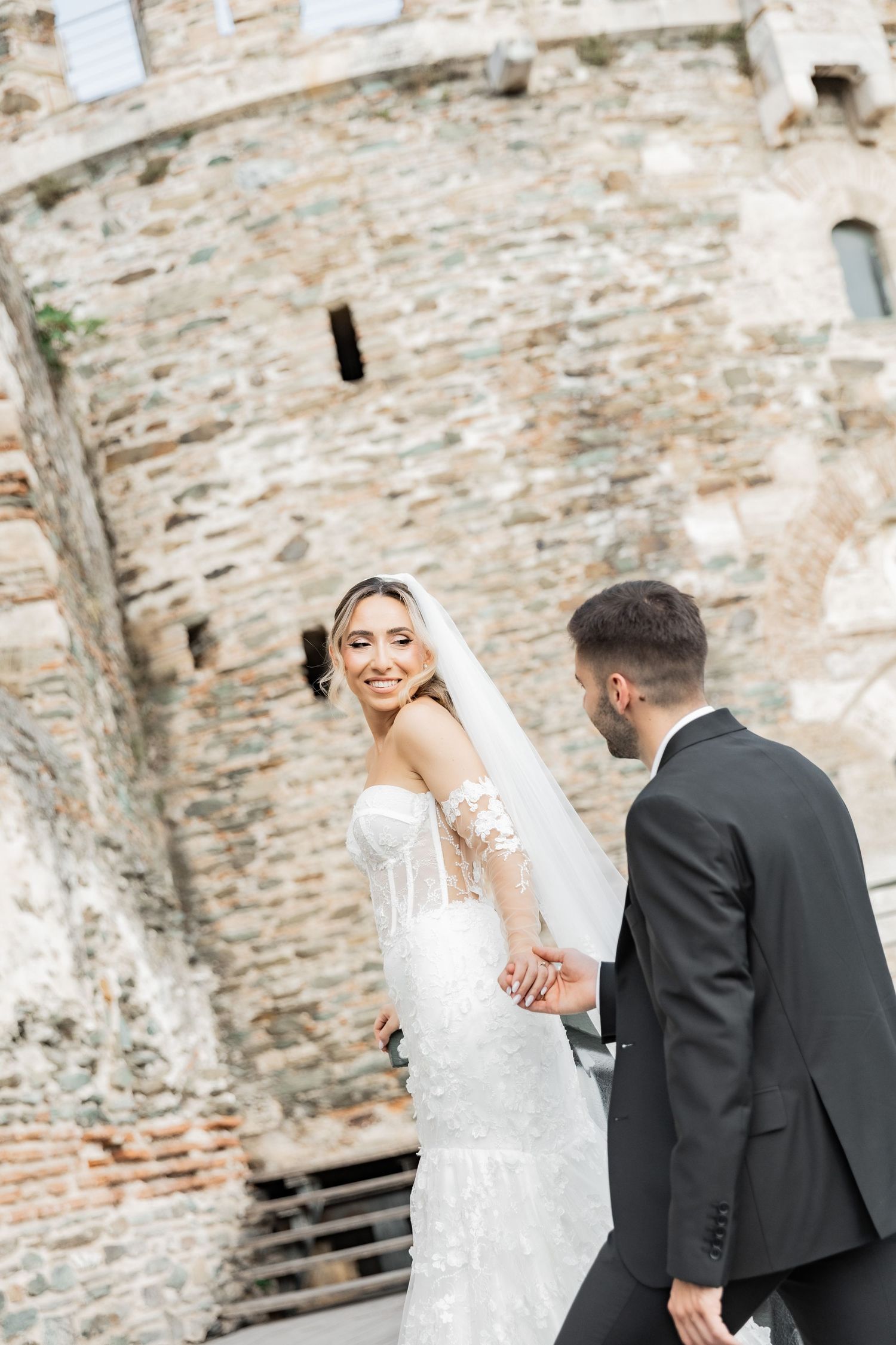 Bride in white lace gown and veil holds grooms hand near historic stone castle walls during outdoor wedding photoshoot.
