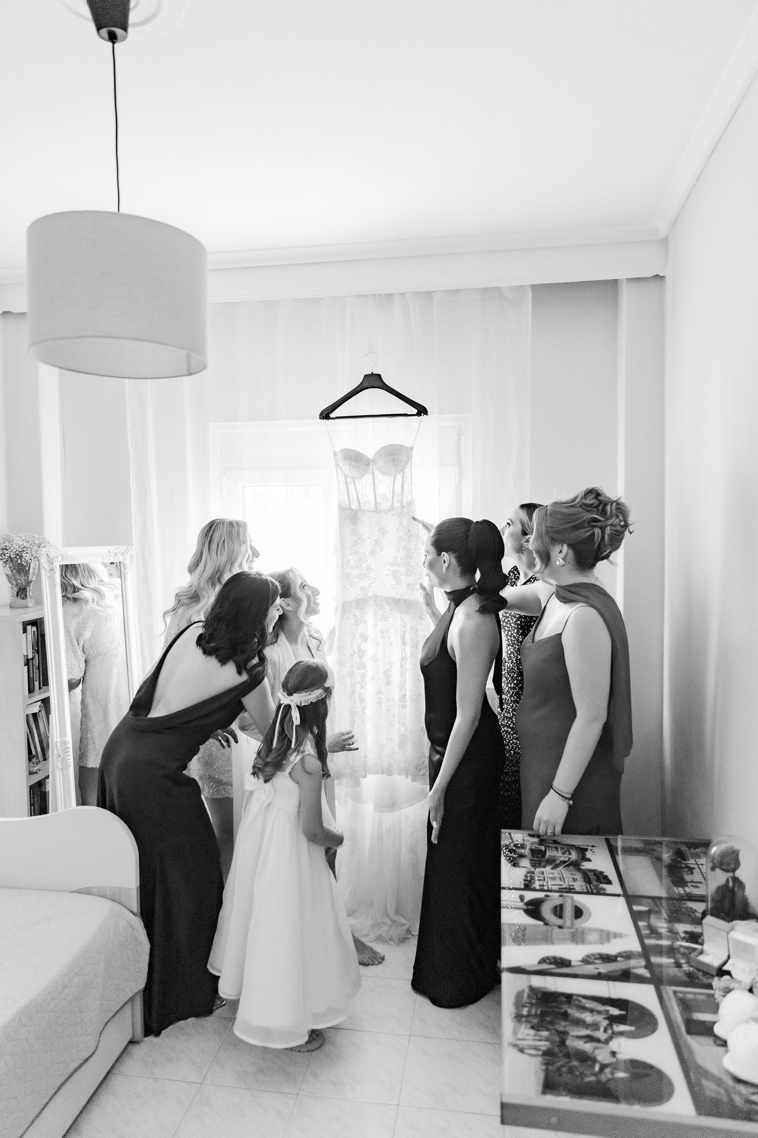 Black and white photo of bridesmaids admiring a wedding dress hanging by a window during bridal preparations.