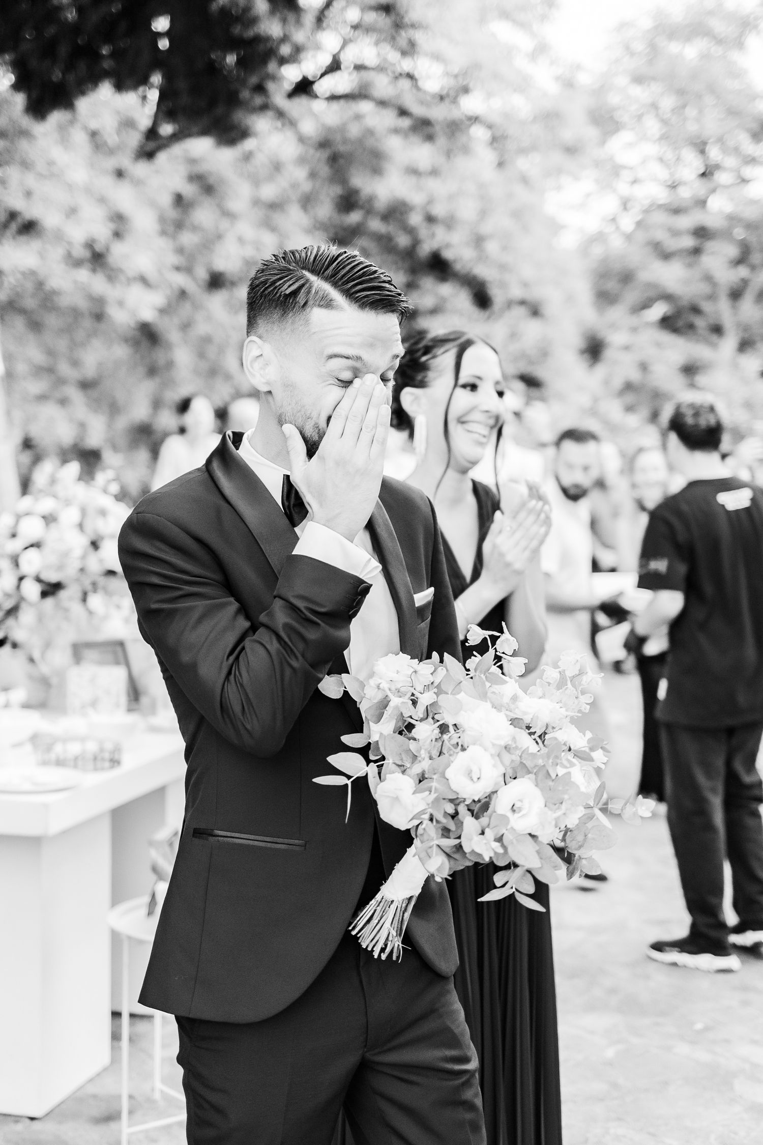 Black and white photo of an emotional wedding moment with bouquets and formal attire.