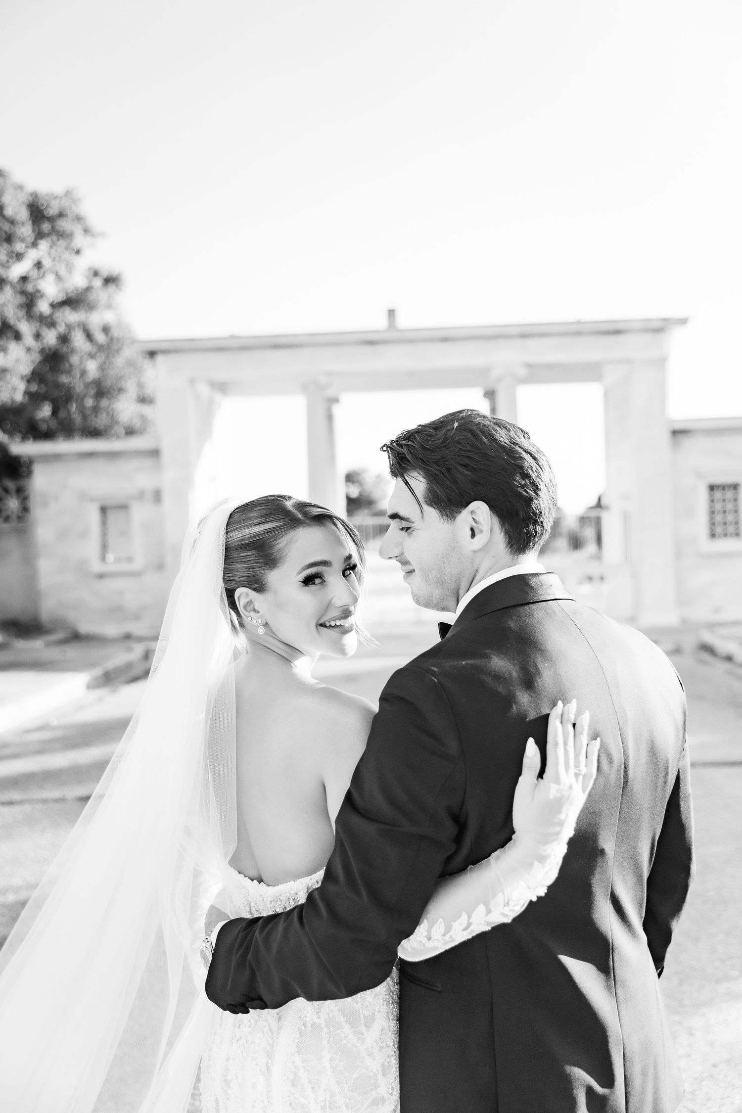Black and white photo of a smiling bride in a veil and groom in a suit embracing outdoors near a classical building.