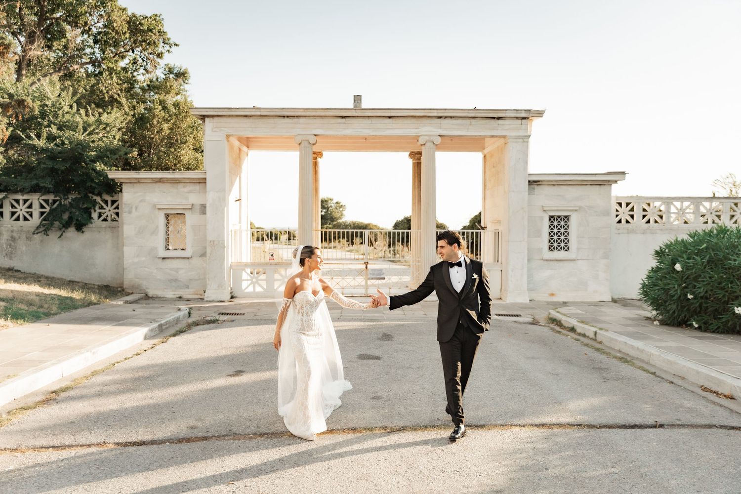 Bride and groom hold hands walking in front of elegant white stone columns at an outdoor wedding venue.