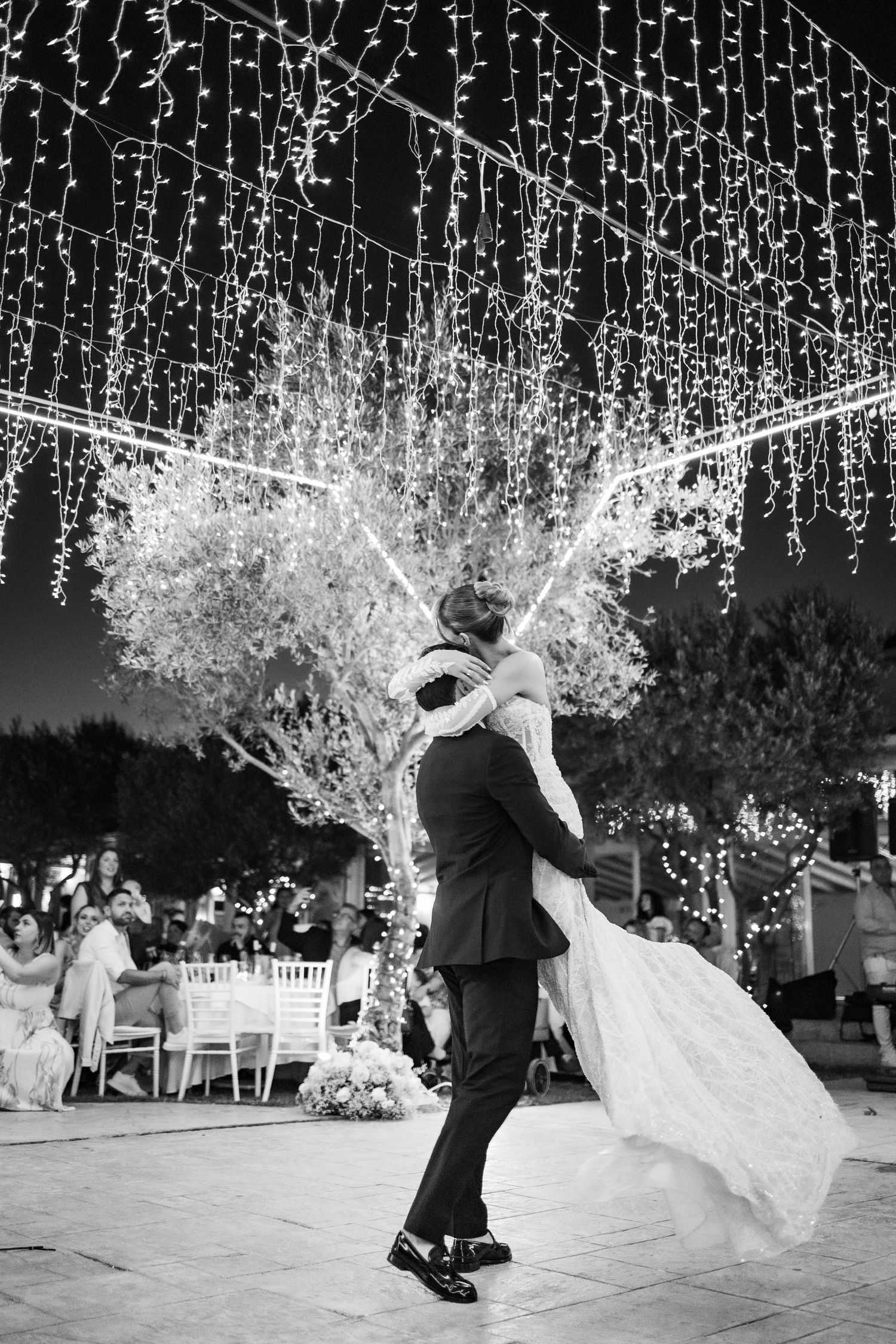 Black and white photo of a groom lifting his bride during their first dance under twinkling fairy lights outdoors.
