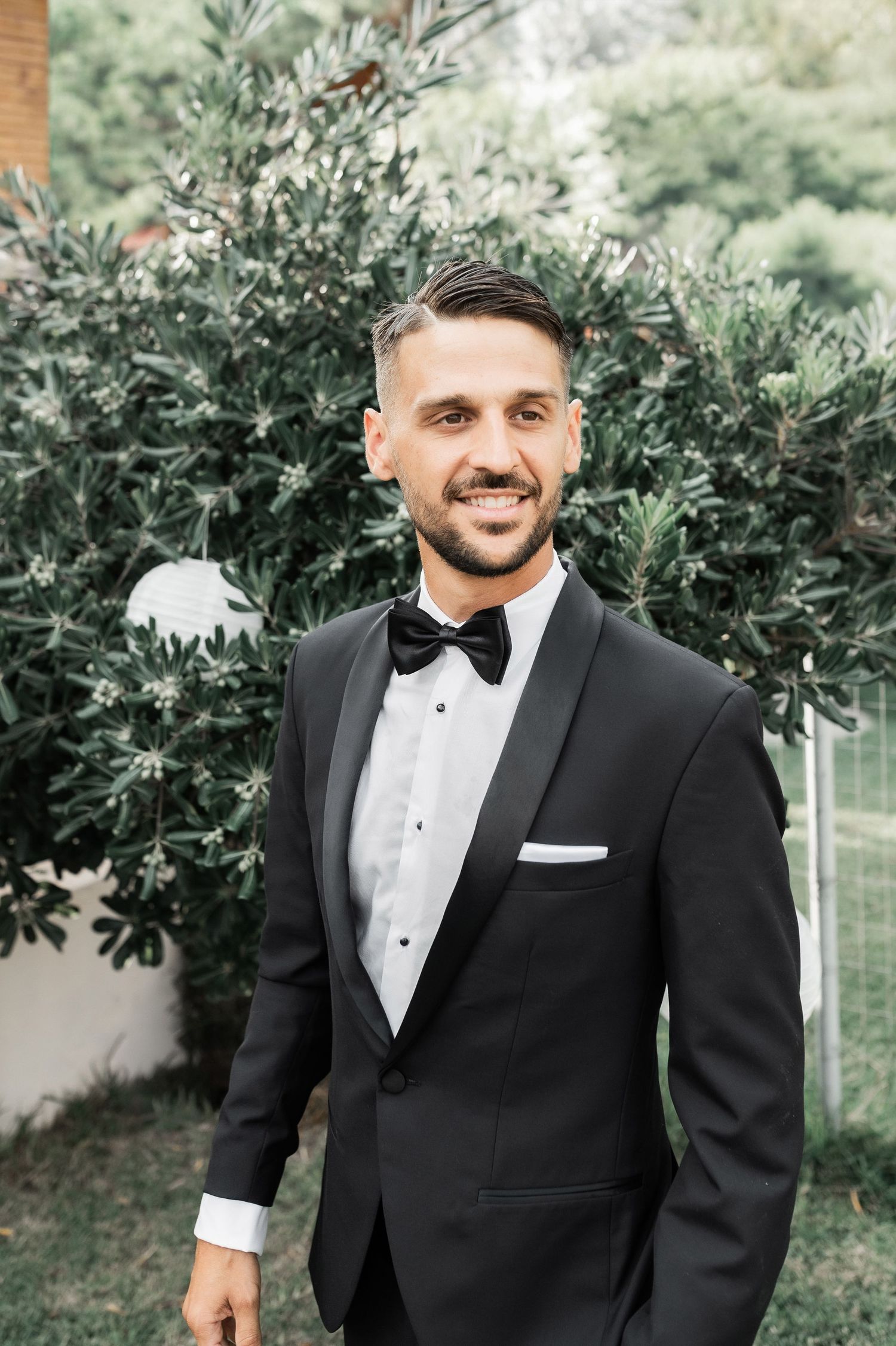 Groom in classic black tuxedo poses in garden setting.