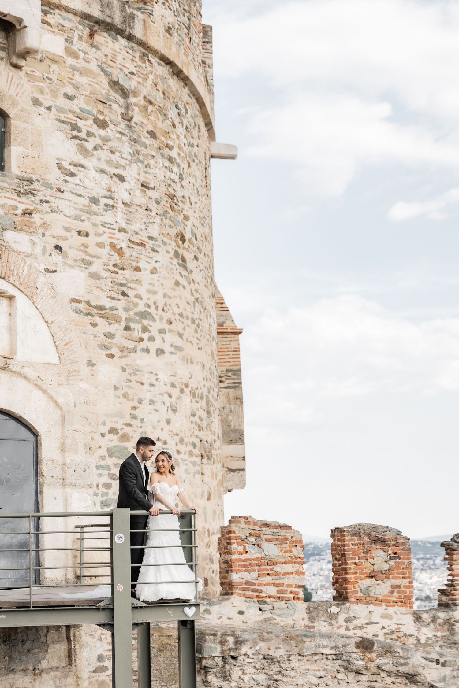 Bride and groom posing on a balcony of a historic stone castle tower with scenic ruins in the background.