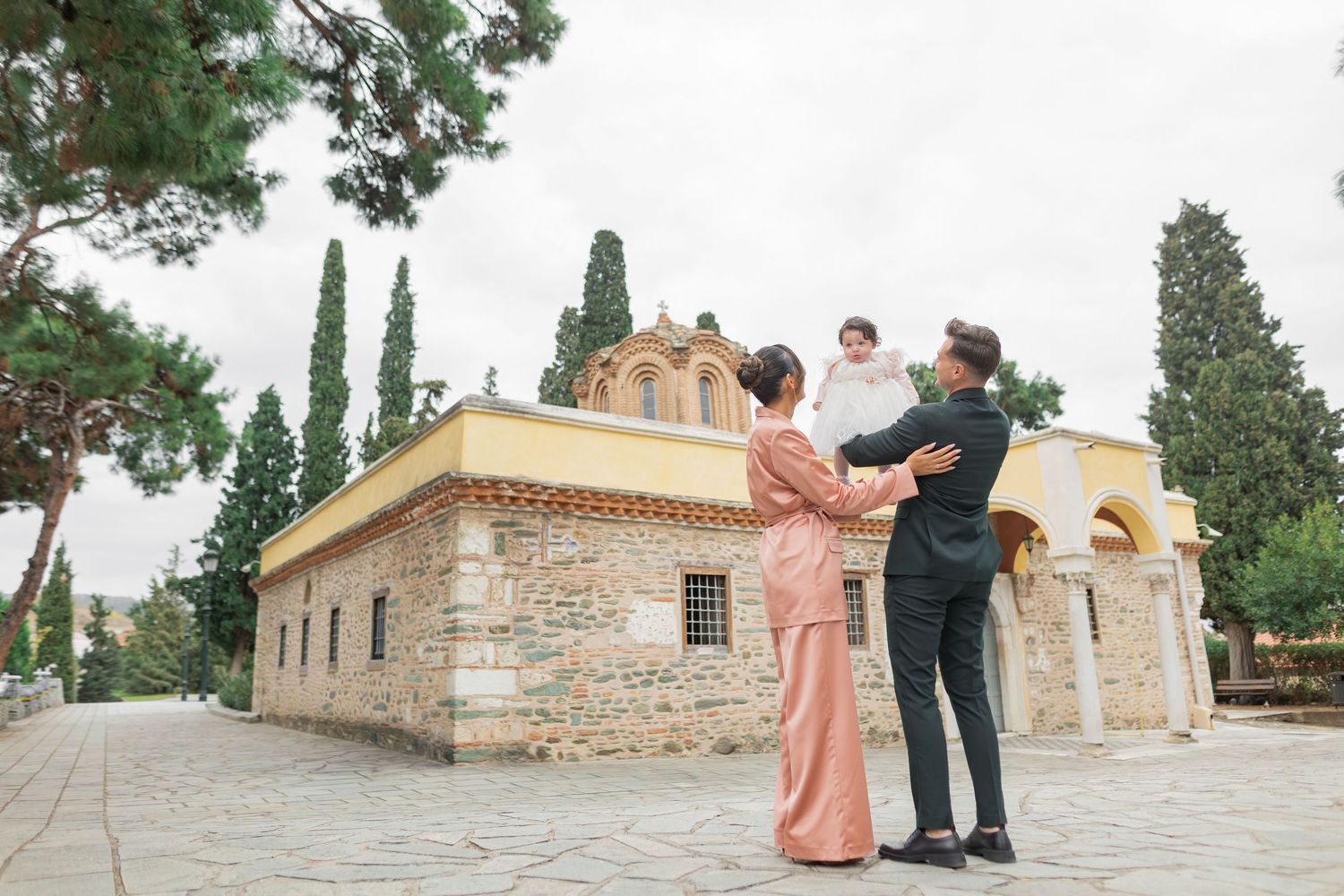 A couple dances together outside a historic stone church with cypress trees in Greece.
