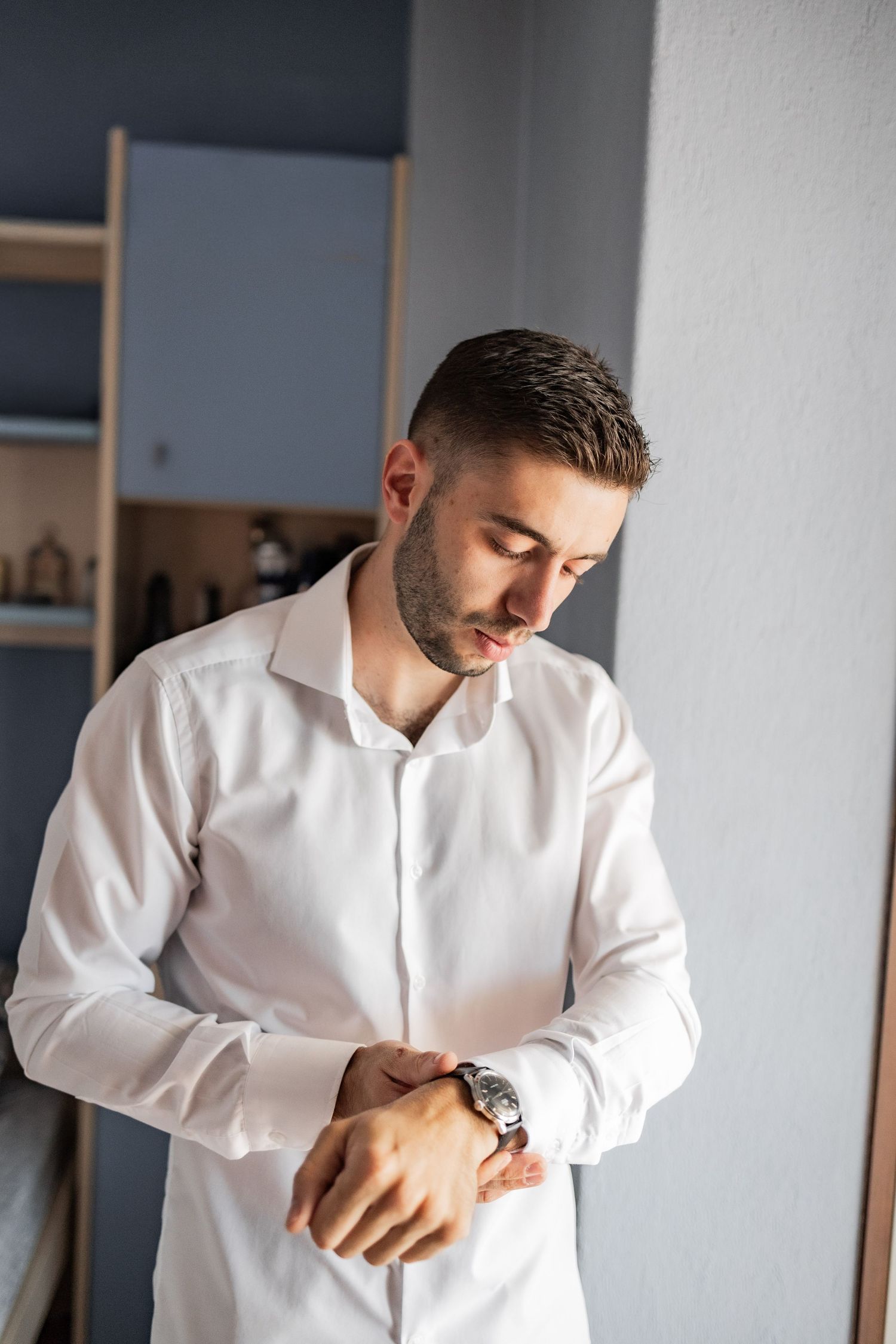 Young man in white dress shirt adjusting his watch cufflink while getting dressed indoors.
