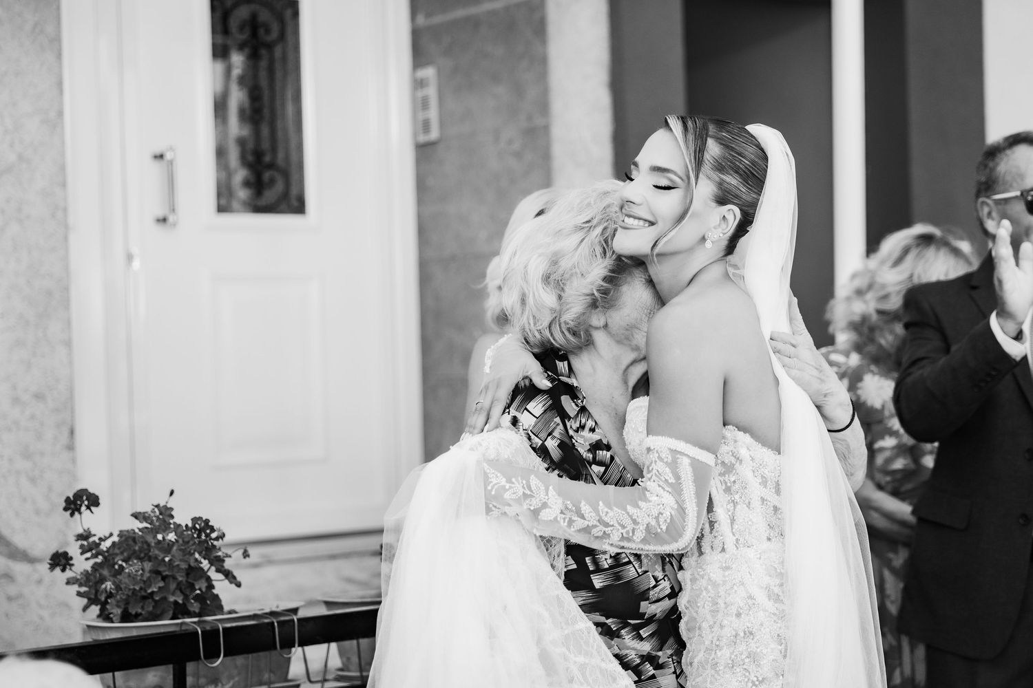Black and white photo of a bride receiving a warm hug and kiss from an older woman at a wedding celebration.