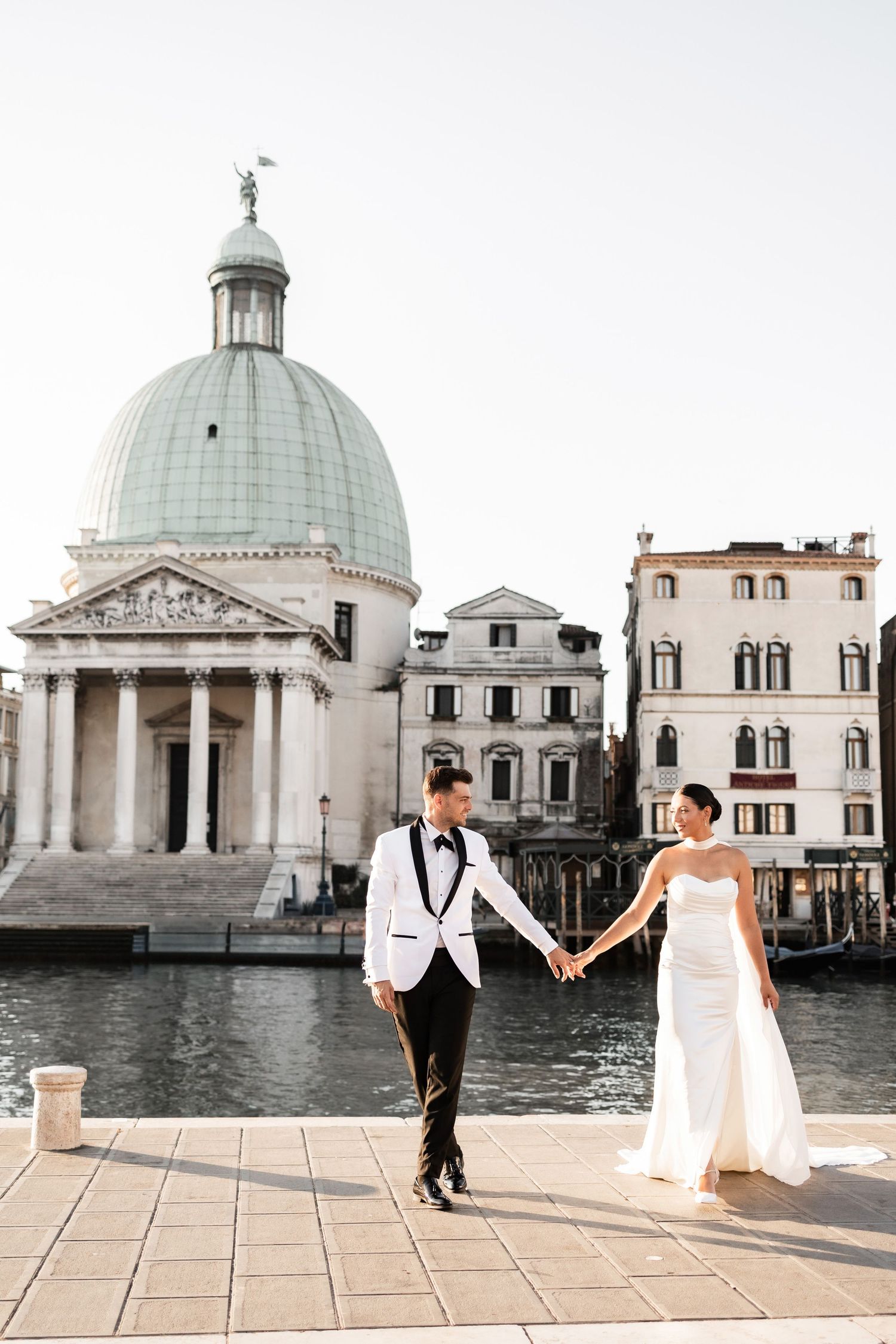 Wedding couple walking along Venice's waterfront with historic domed buildings in background.