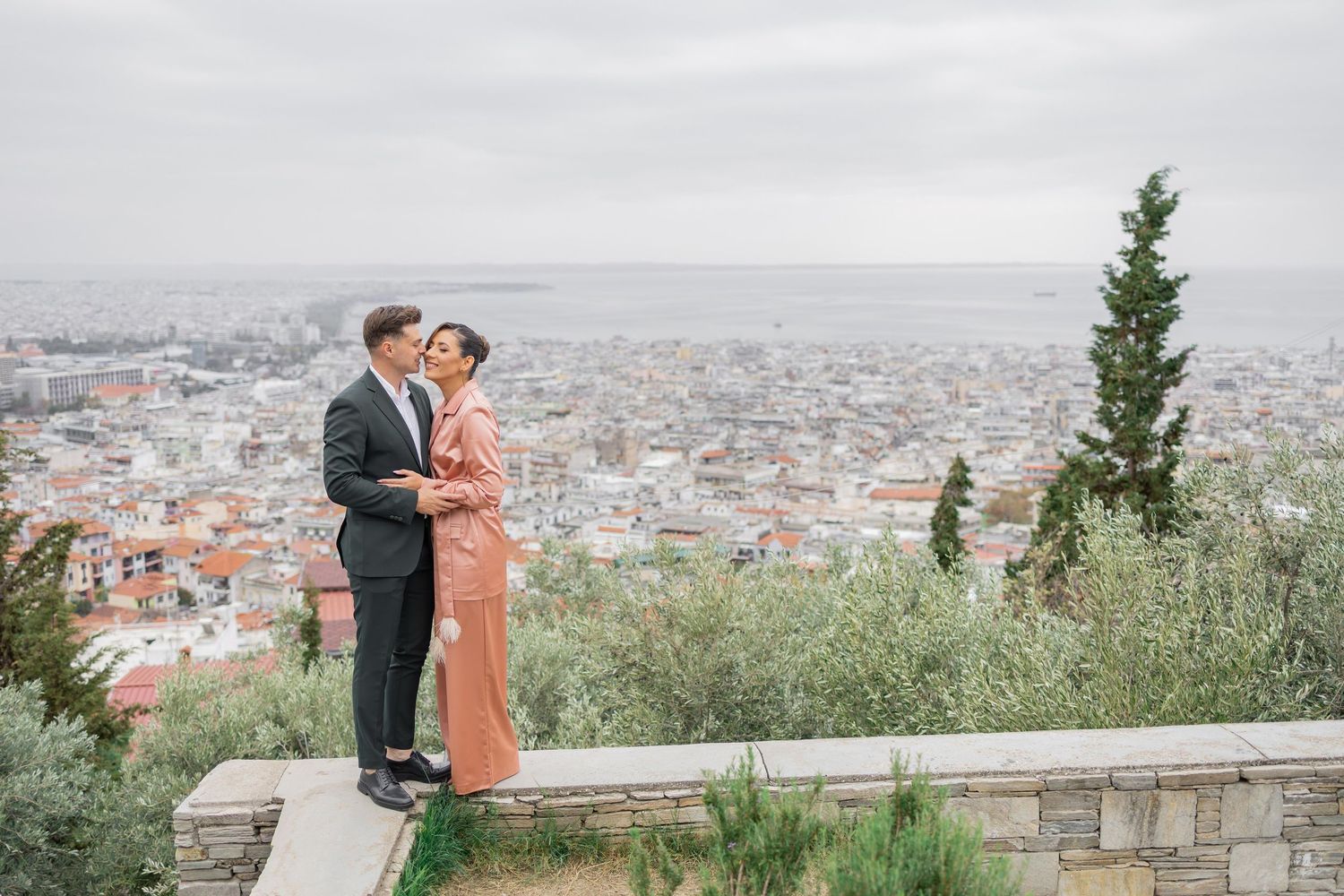 A couple shares a romantic moment overlooking a scenic cityscape view from a hilltop terrace.