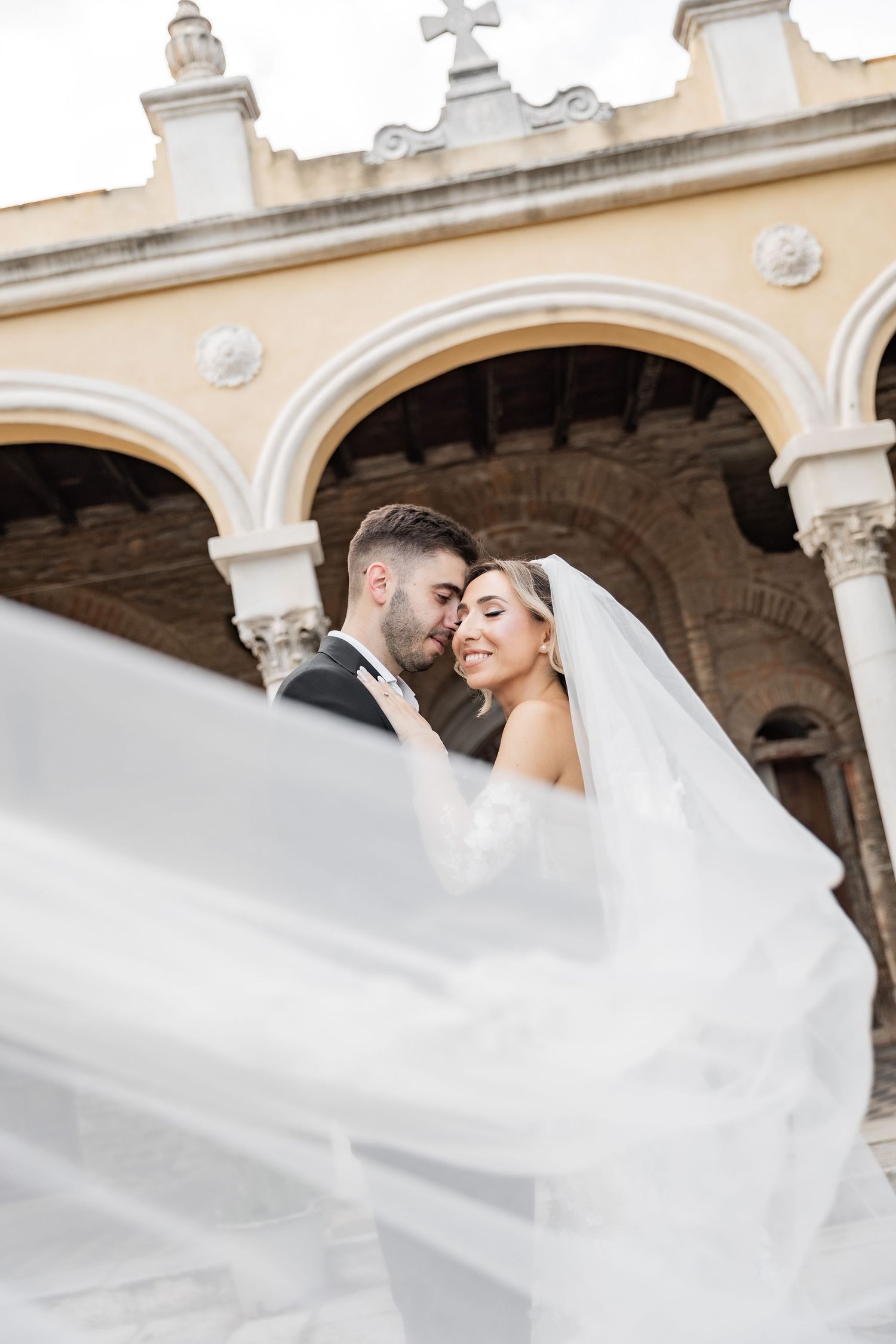Bride and groom embrace in front of elegant arched building, her veil flowing in the wind on their wedding day.