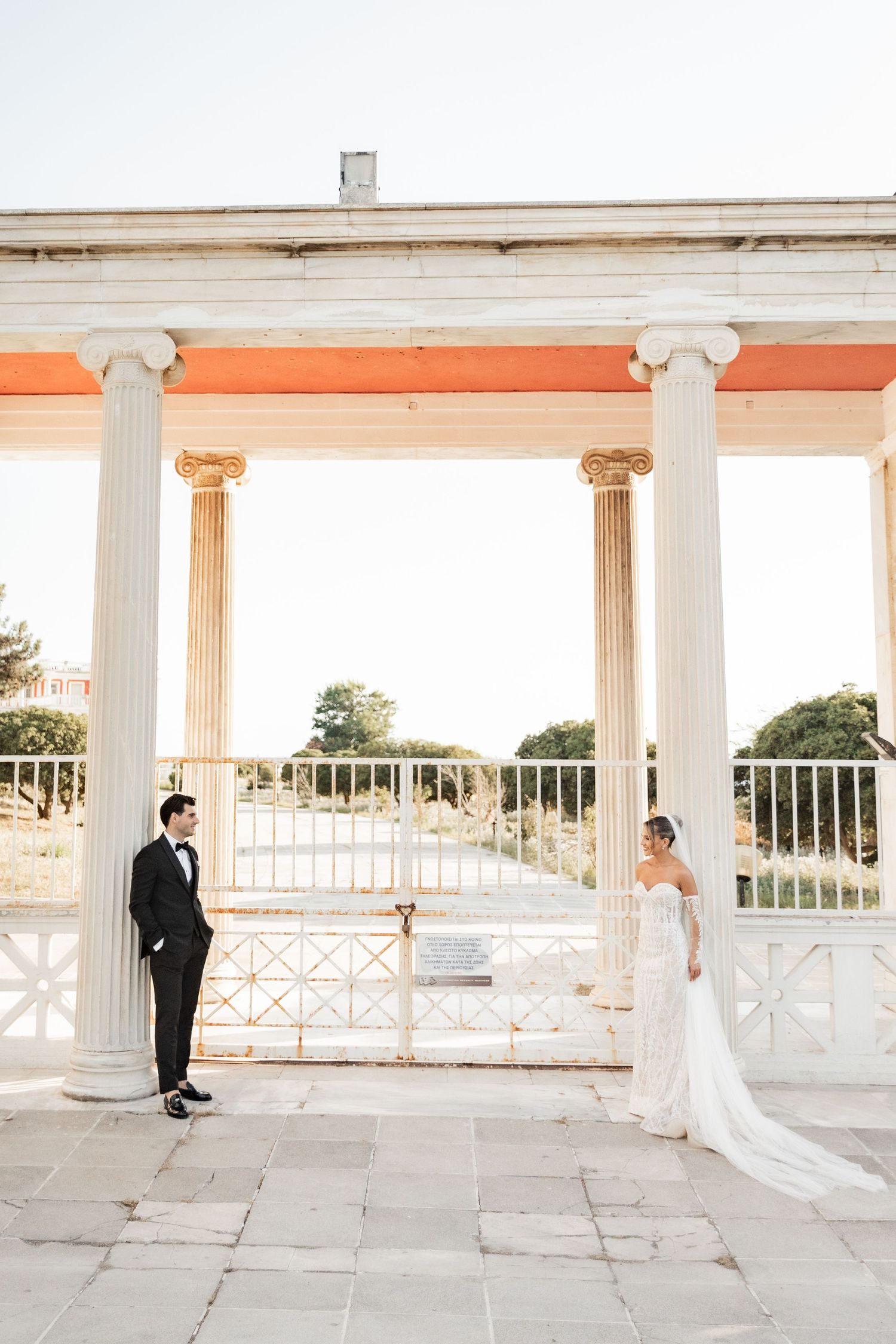 Bride and groom stand apart beneath white Greek columns at an elegant outdoor wedding venue with iron gates.