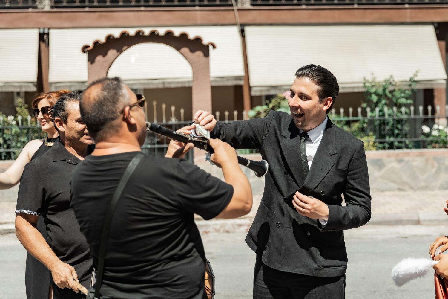 Man in suit gestures while speaking to group outdoors on sunny day near ornate building.