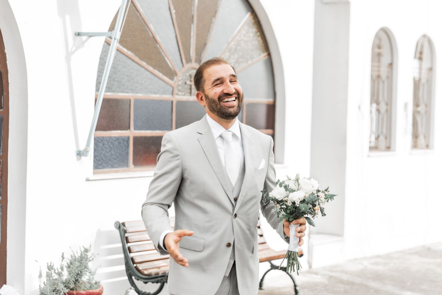 Wedding party members in light gray suits share laughs in front of white archways.