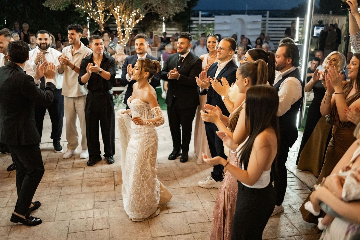 Bride and groom share first dance surrounded by clapping wedding guests at elegant outdoor evening reception.