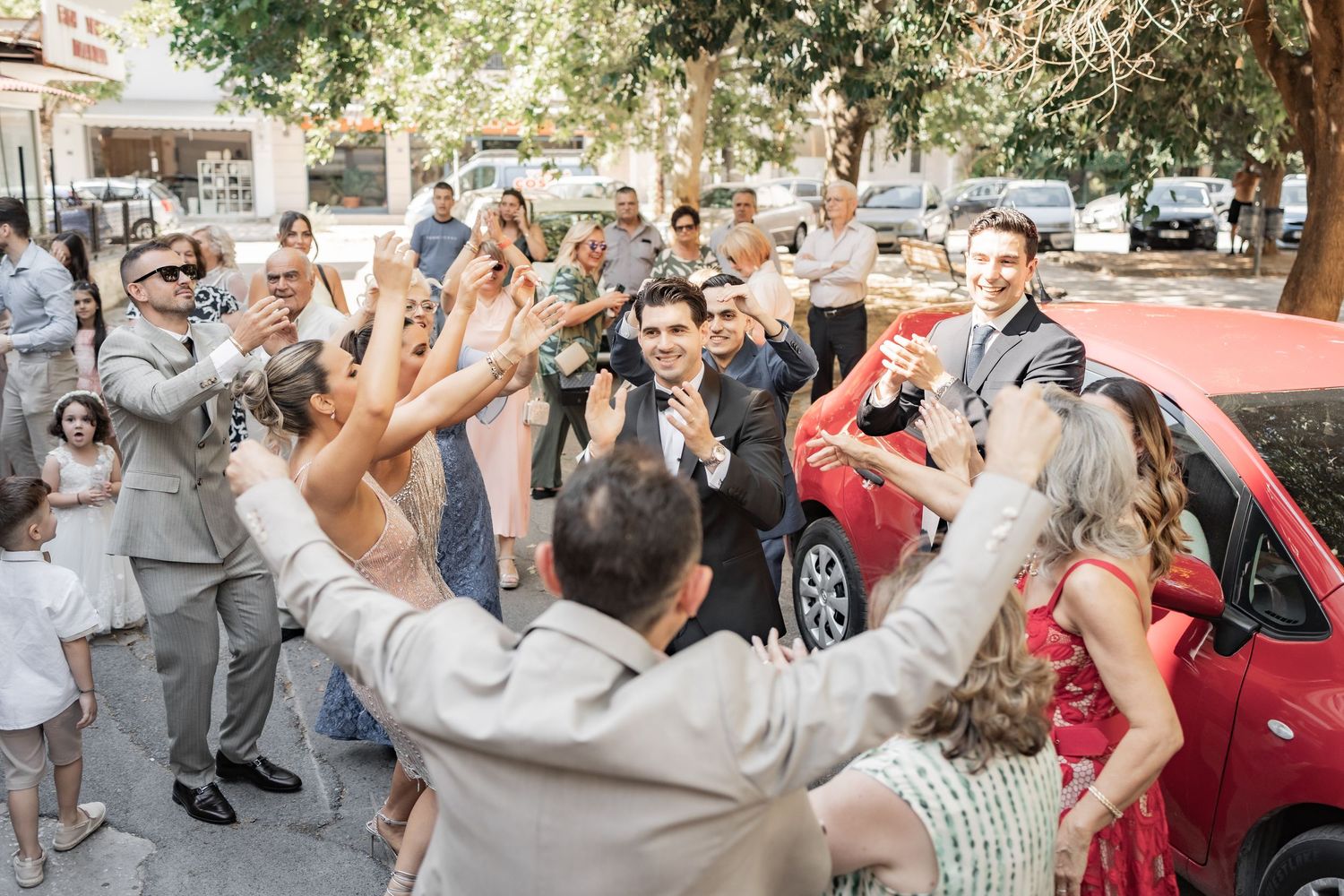 Joyful wedding guests celebrate outdoors, clapping and cheering around the couple near a red car on a sunny day.