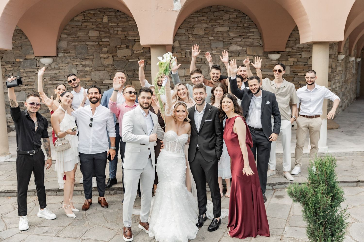 Bride and groom celebrate with wedding guests outdoors at a stone venue, guests cheering with raised arms.