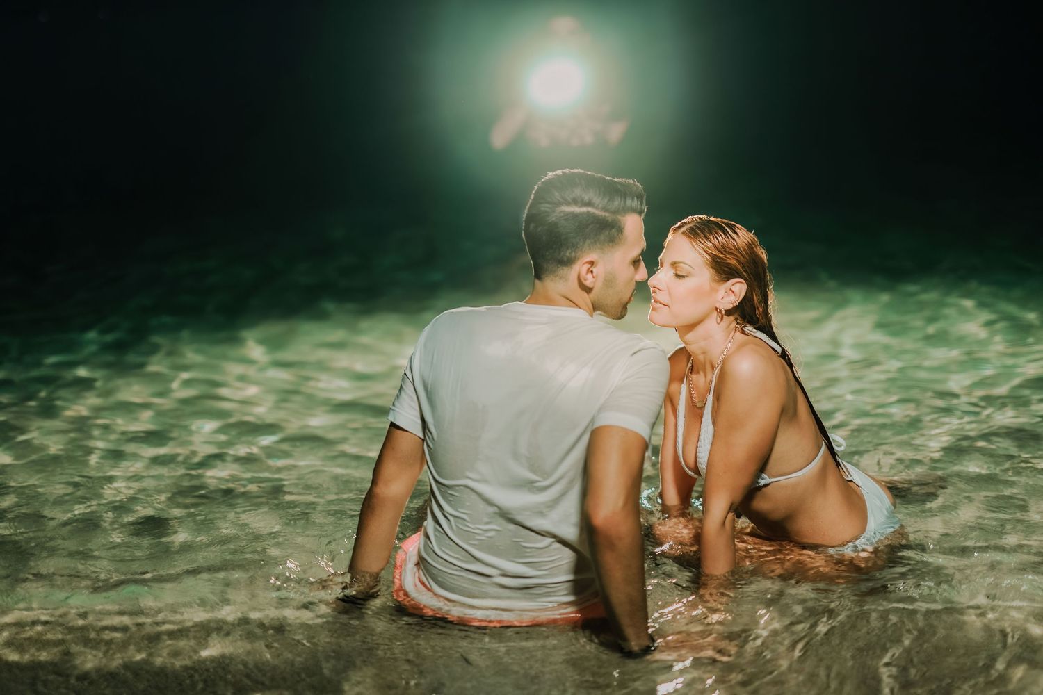 Couple embraces on a moonlit beach with glowing turquoise water in the background.