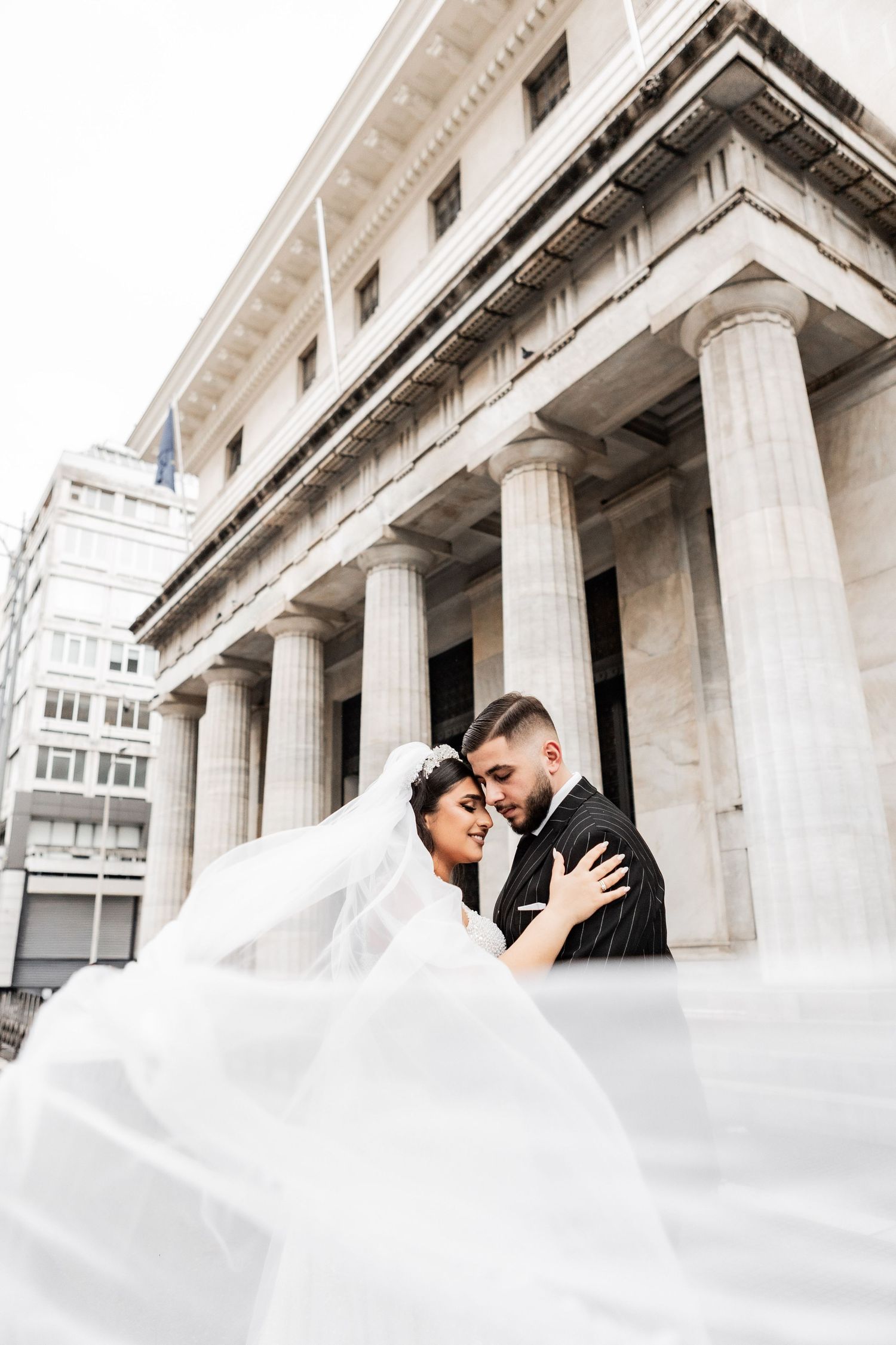 Dynamic wedding photo with flowing veil against classical columned building facade.
