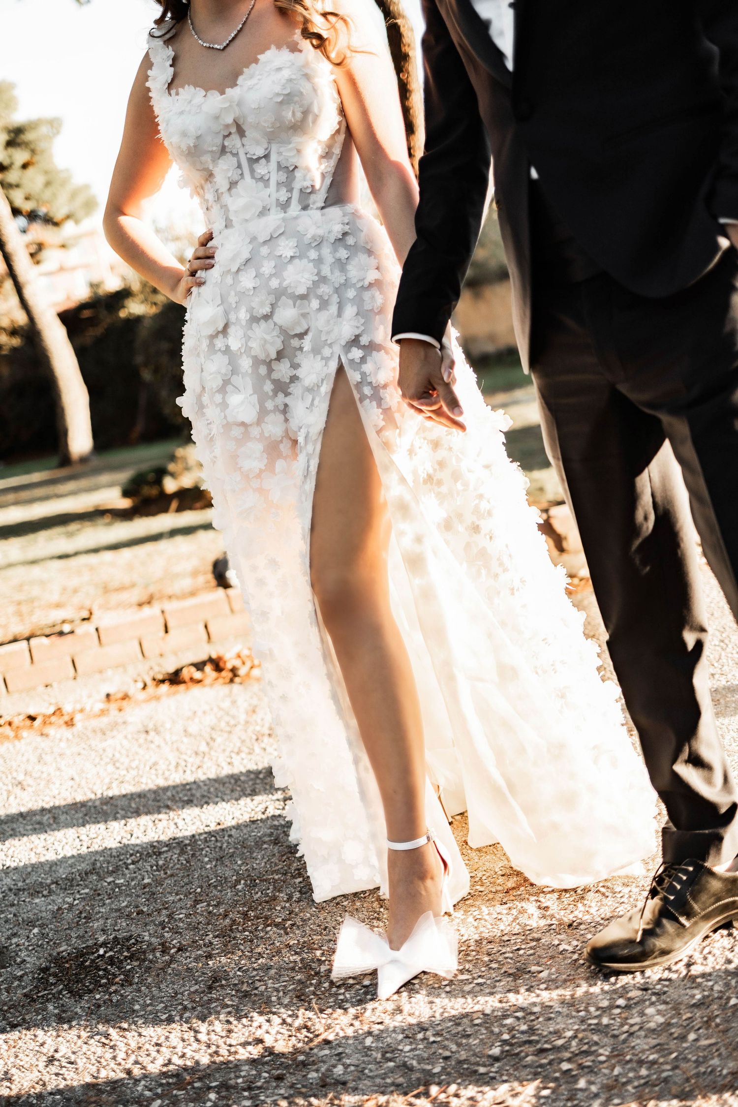 Close-up details of a white floral wedding dress and formal attire on a gravel path.