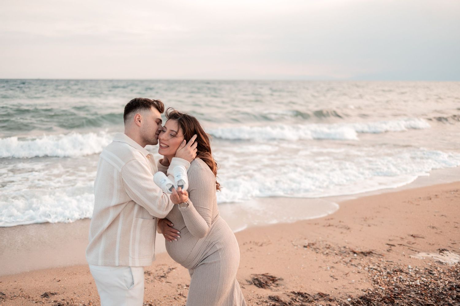 A couple shares a tender embrace on a sandy beach at sunset with ocean waves rolling in the background.