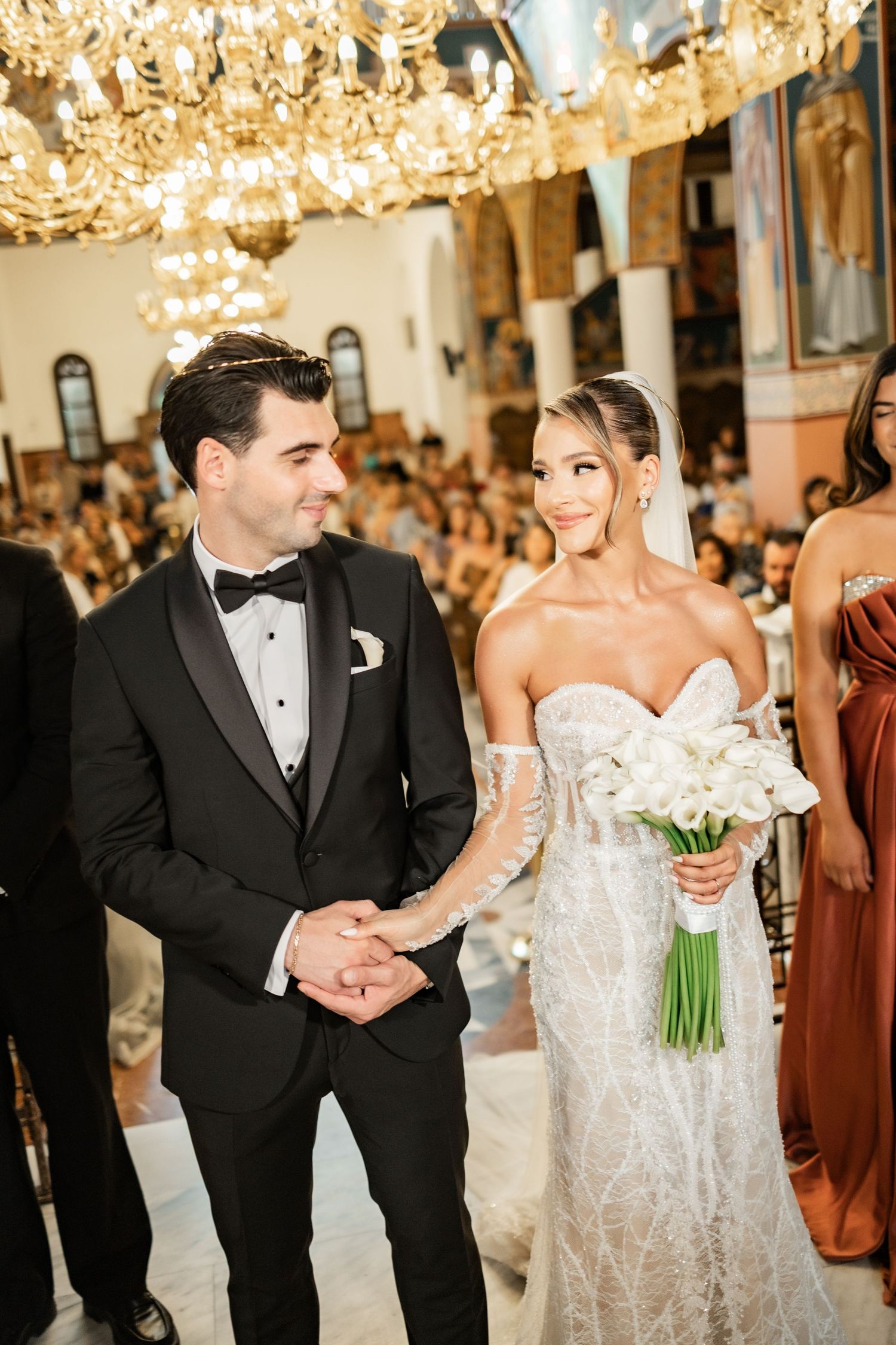 Bride in lace gown holding white bouquet stands with groom in black tuxedo during elegant church wedding ceremony.