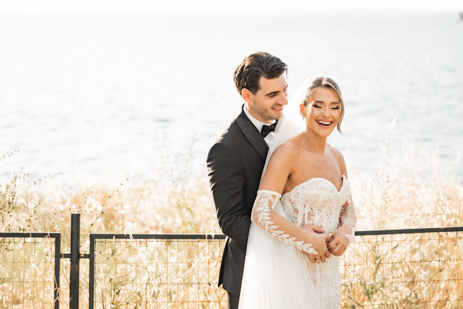 Bride and groom embracing outdoors near waterfront, she in lace wedding gown, he in black tuxedo, golden grass background.