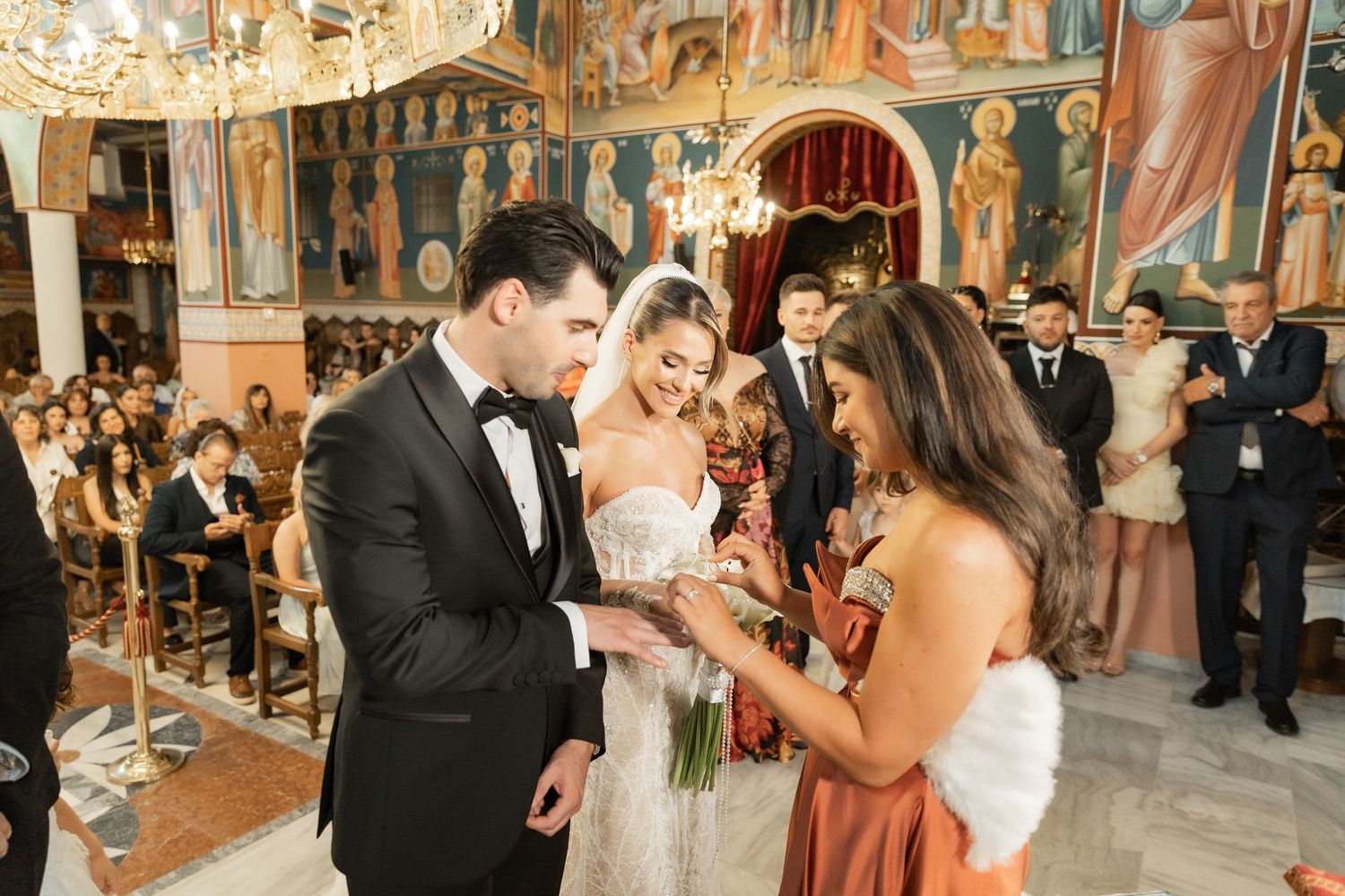 Couple exchanging rings during an Orthodox church wedding ceremony with bridesmaids and guests in ornate decorated interior.