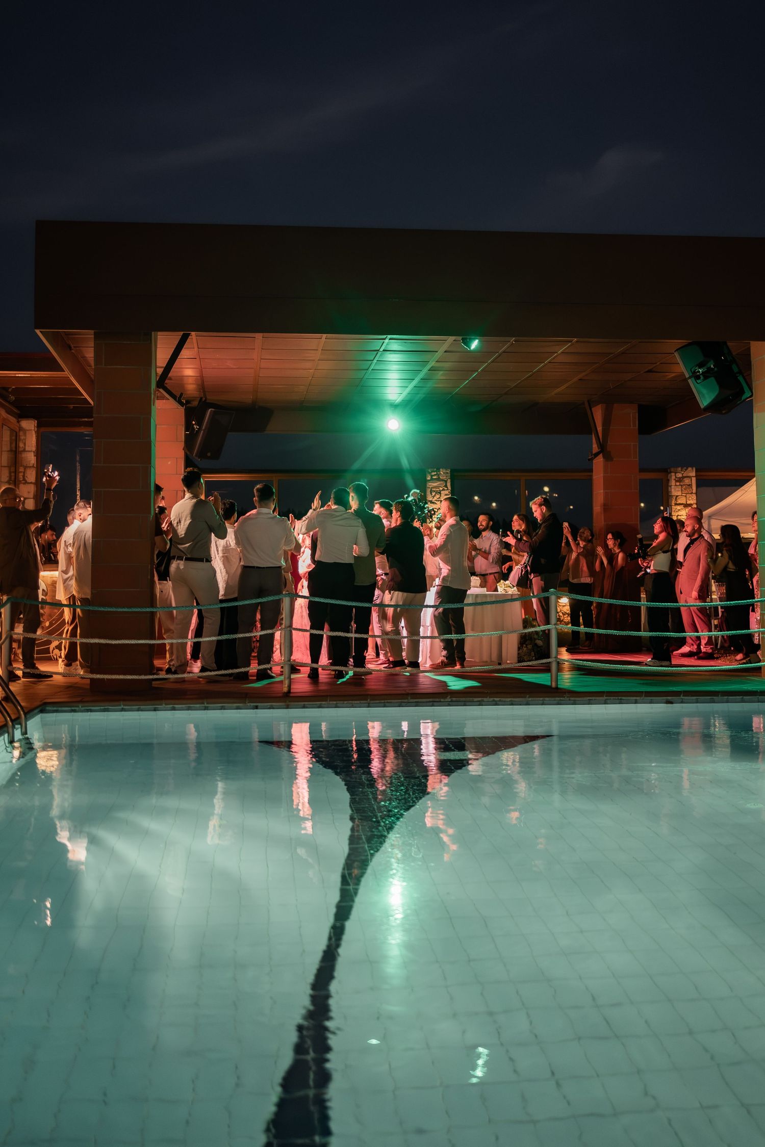 Nighttime pool party with crowd gathered near illuminated stage reflecting green lights on calm water surface.
