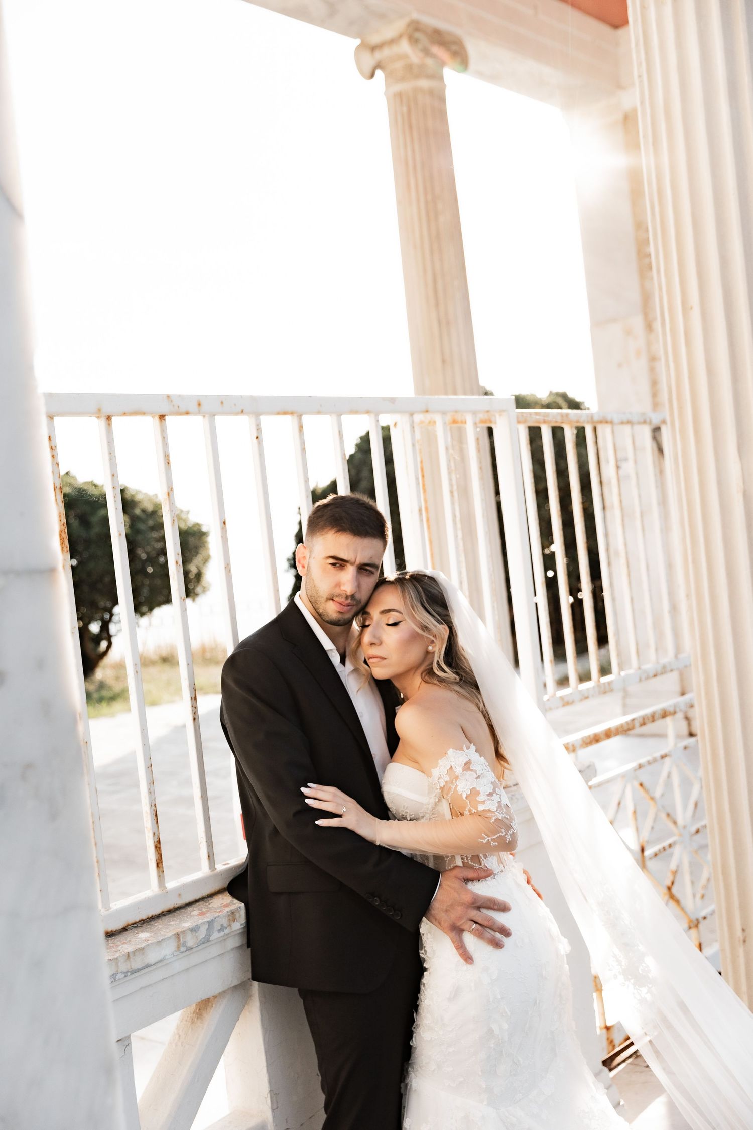 A bride and groom embrace on a sunlit marble balcony with columns, the bride in a lace gown and veil.