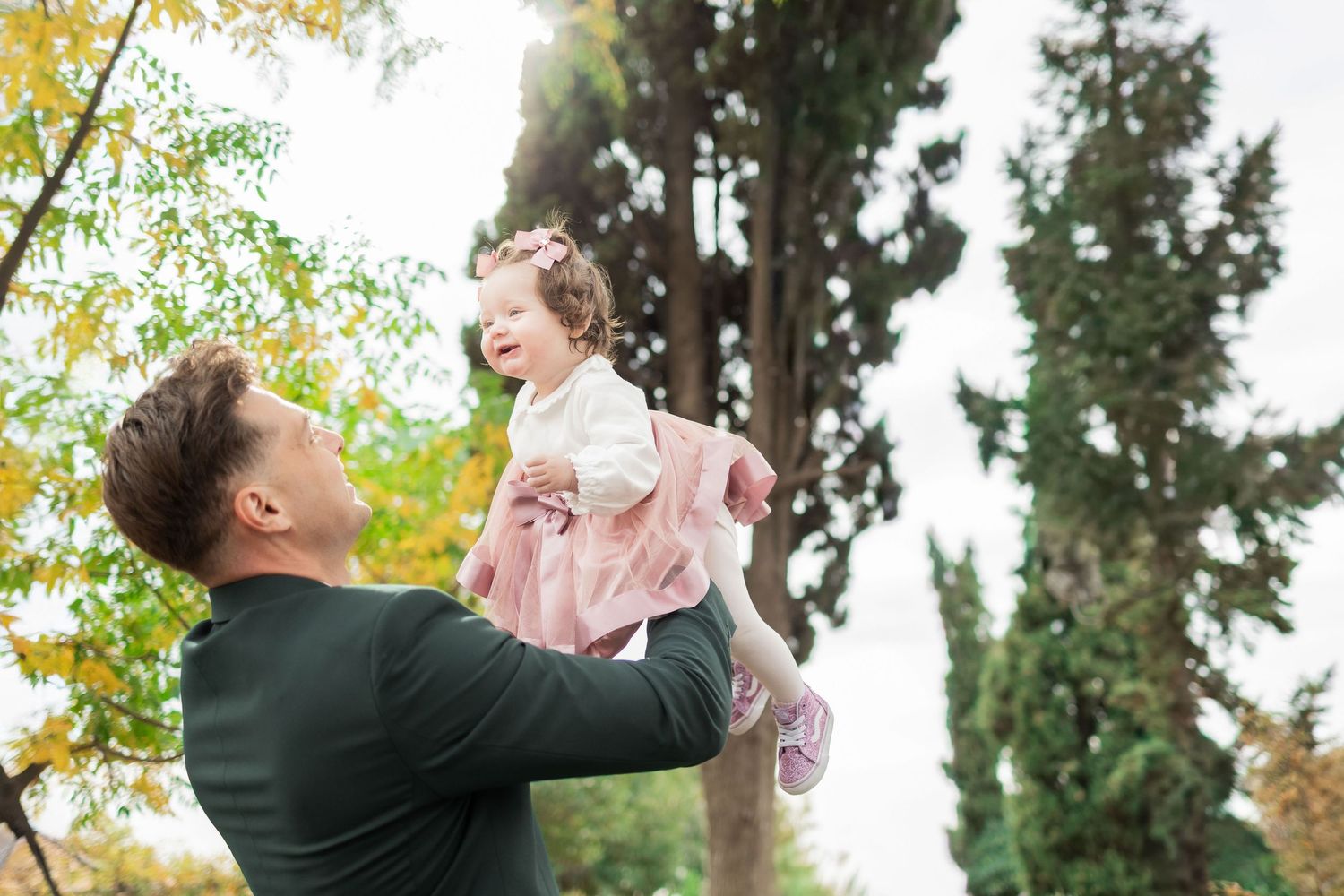 A parent lifts a small child wearing a pink sweater up into the air among autumn trees on a bright day.